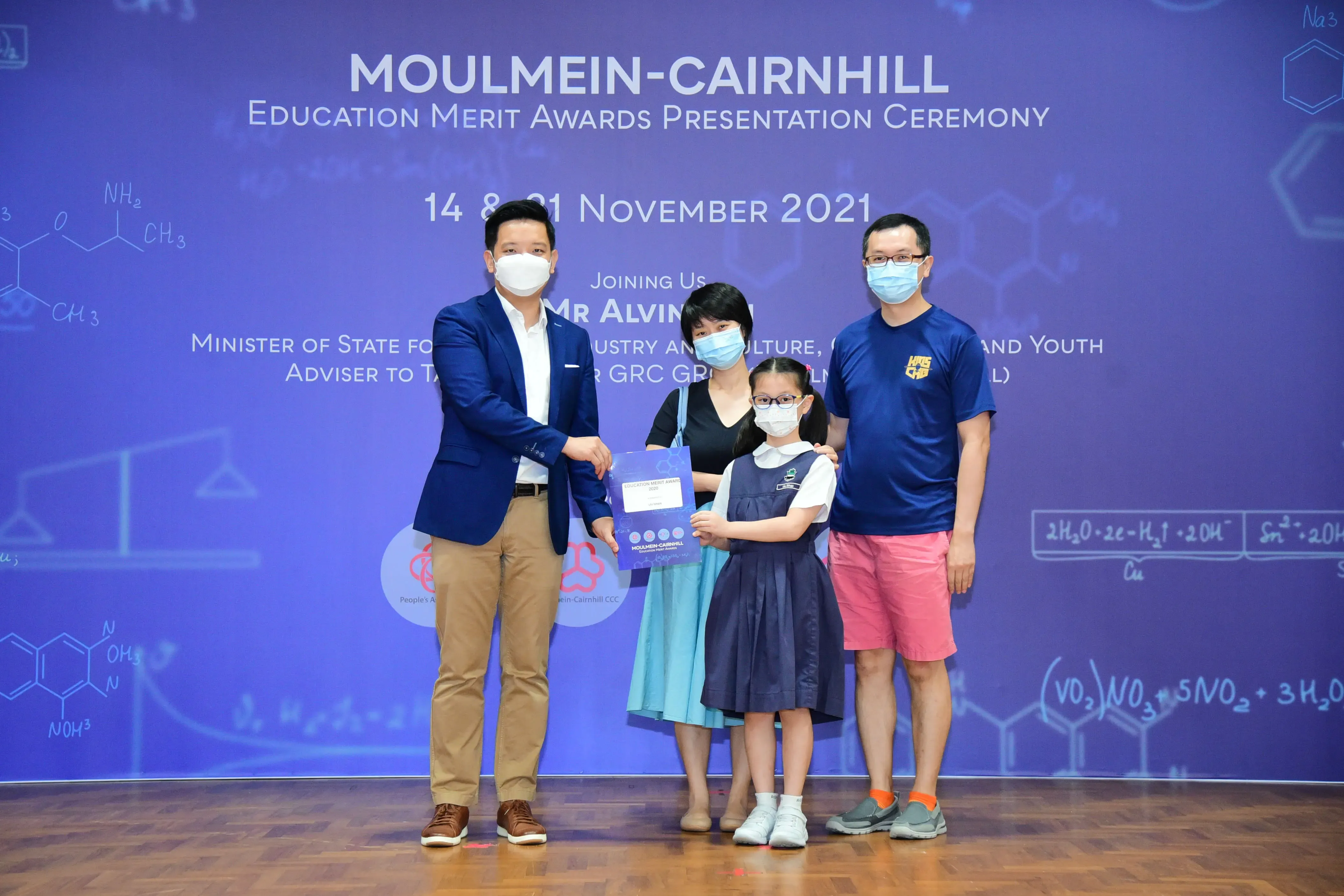 Four masked people pose at the Moulmein-Cairnhill Education Merit Awards Ceremony, November 2021, with a child holding an award.