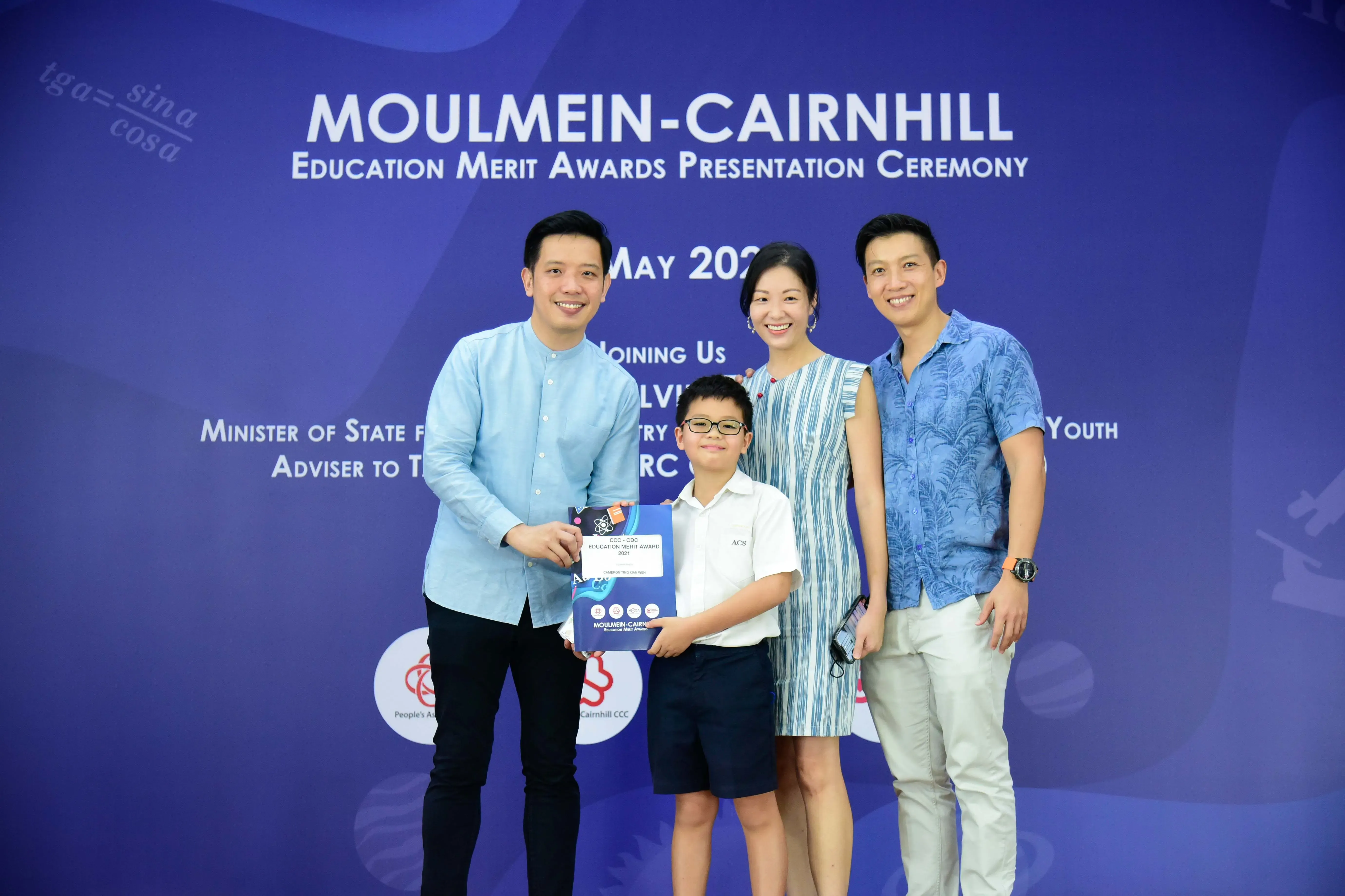 Three adults and a boy holding an award certificate pose in front of a Moulmein-Cairnhill Education Merit Awards banner.
