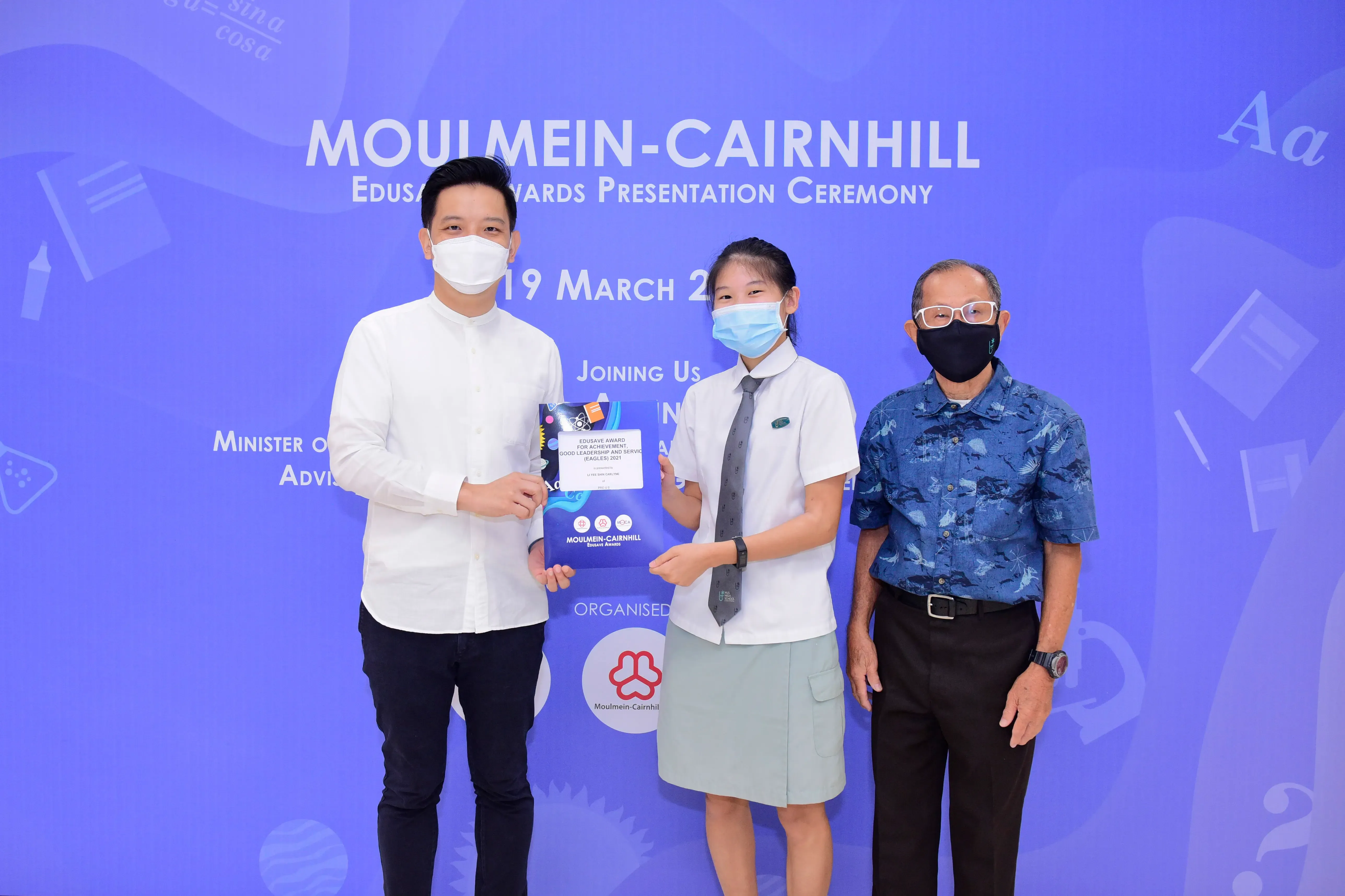 Three masked people pose at the Moulmein-Cairnhill Edusave Awards Presentation Ceremony, with a student holding an award folder.
