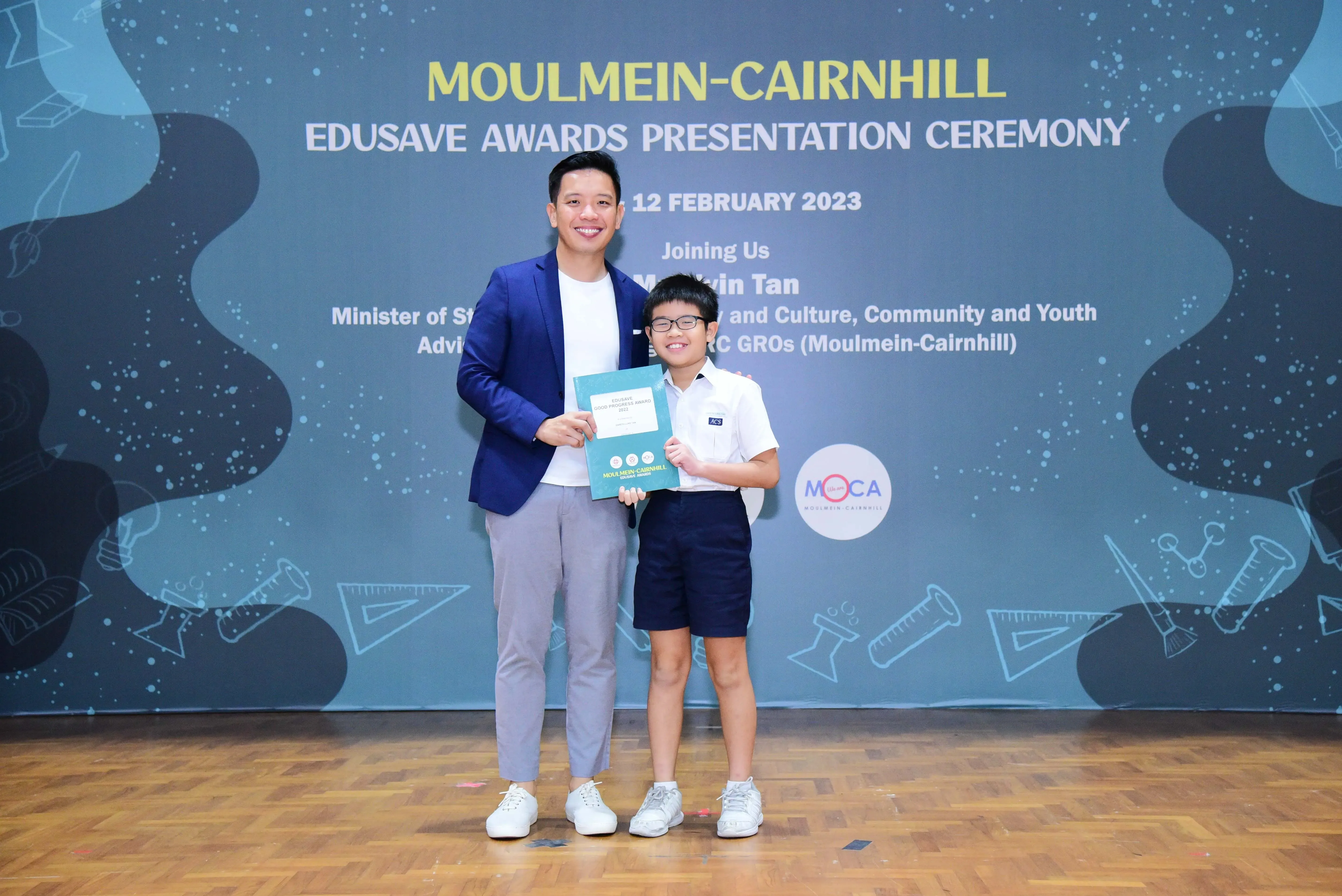 An adult in a blue blazer and a young student in school uniform hold a certificate at the Moulmein-Cairnhill Edusave Awards 2023.