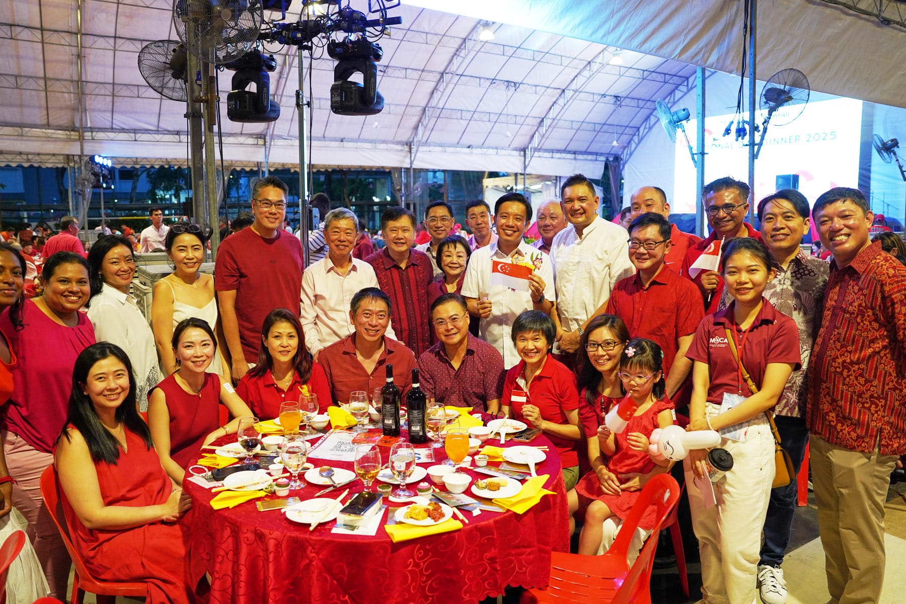 Large group of smiling people in red outfits posing around a dinner table at an outdoor event under a tent.