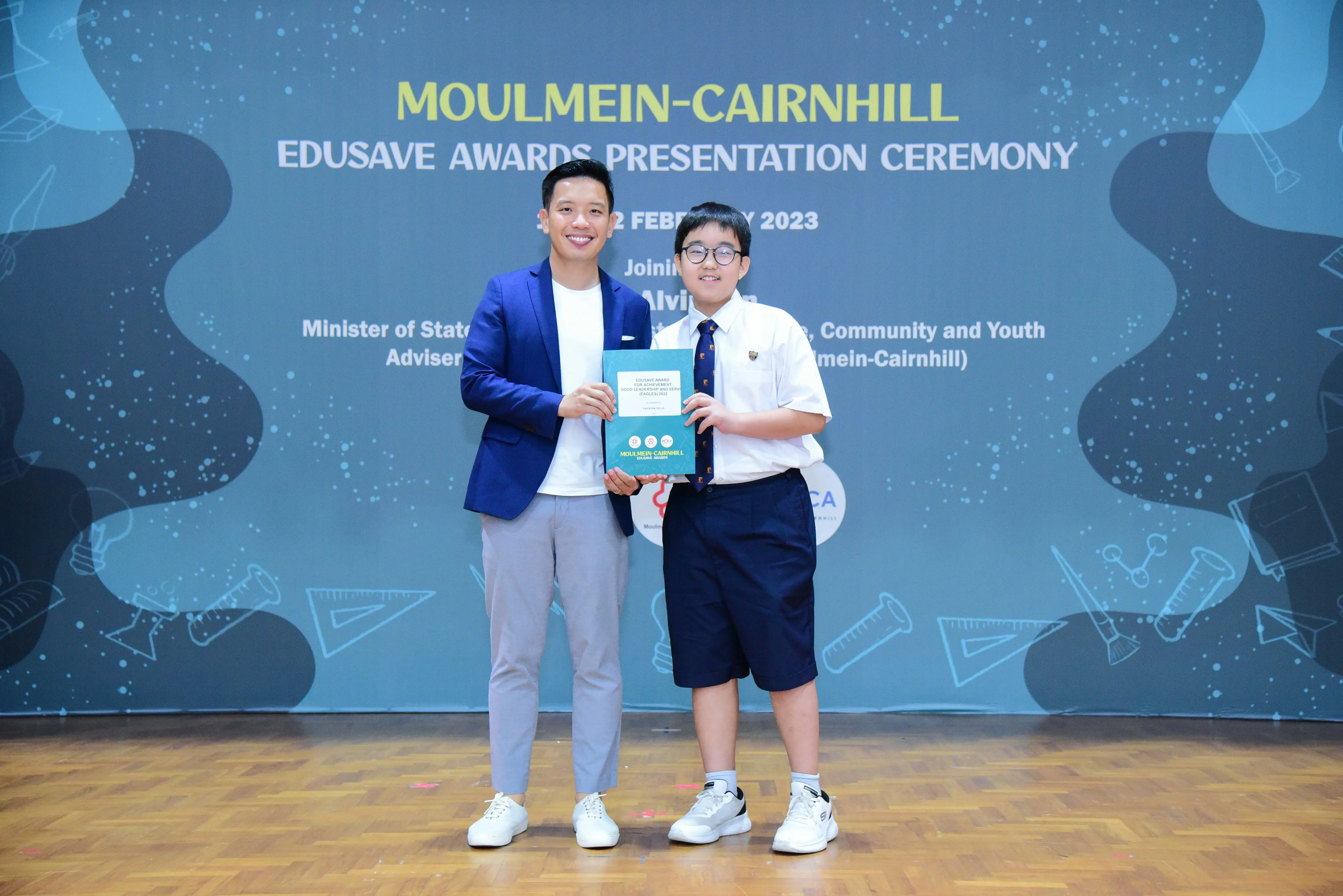 Man in blue blazer and young student in school uniform hold an award together at Moulmein-Cairnhill Edusave ceremony.