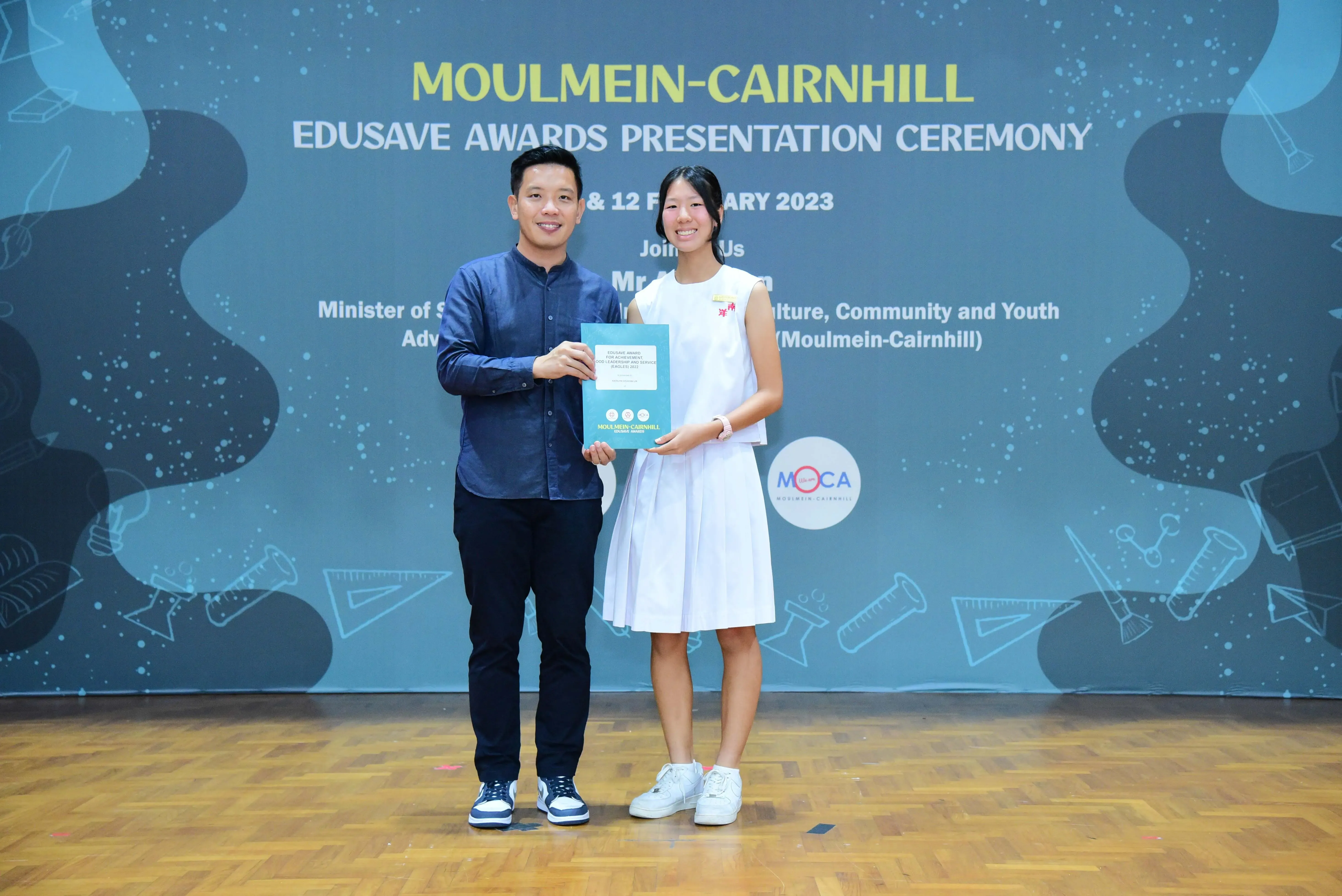 Two people stand on a stage holding a certificate at the Moulmein-Cairnhill Edusave Awards Ceremony 2023.