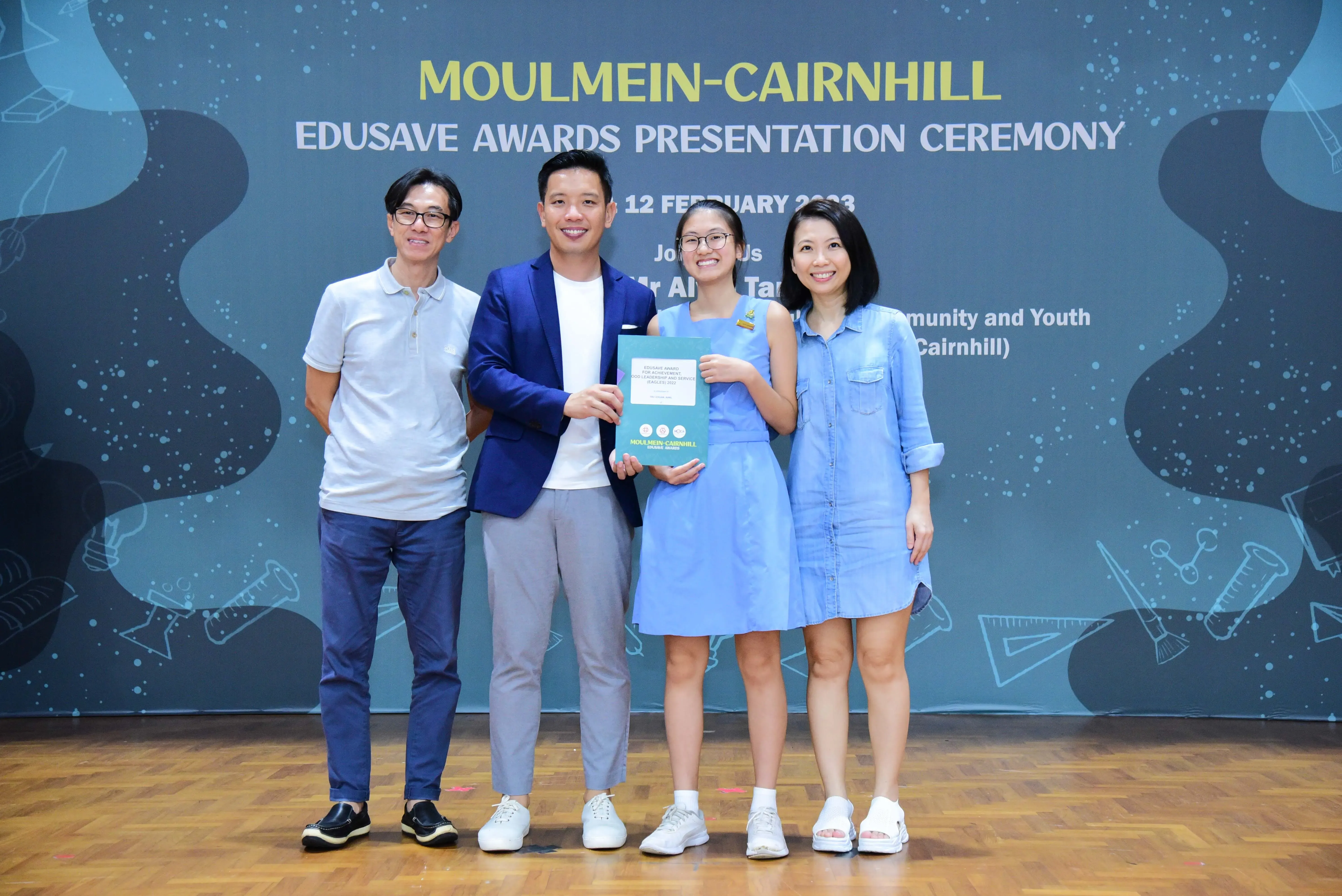 Four smiling people pose at the Moulmein-Cairnhill Edusave Awards Presentation Ceremony, with a girl holding a certificate.