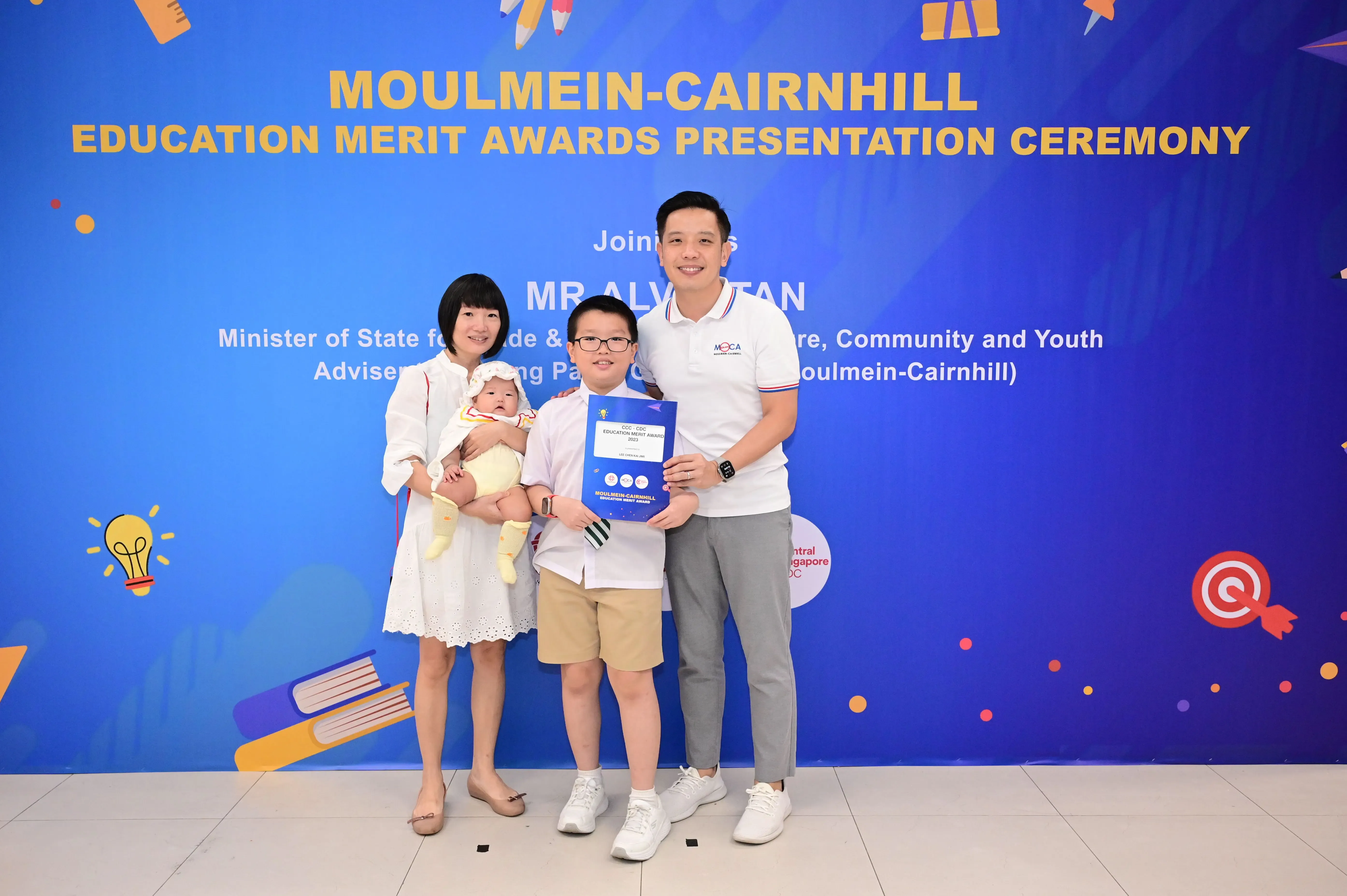 Family of four poses before a Moulmein-Cairnhill Education Merit Awards banner; boy holds a certificate.