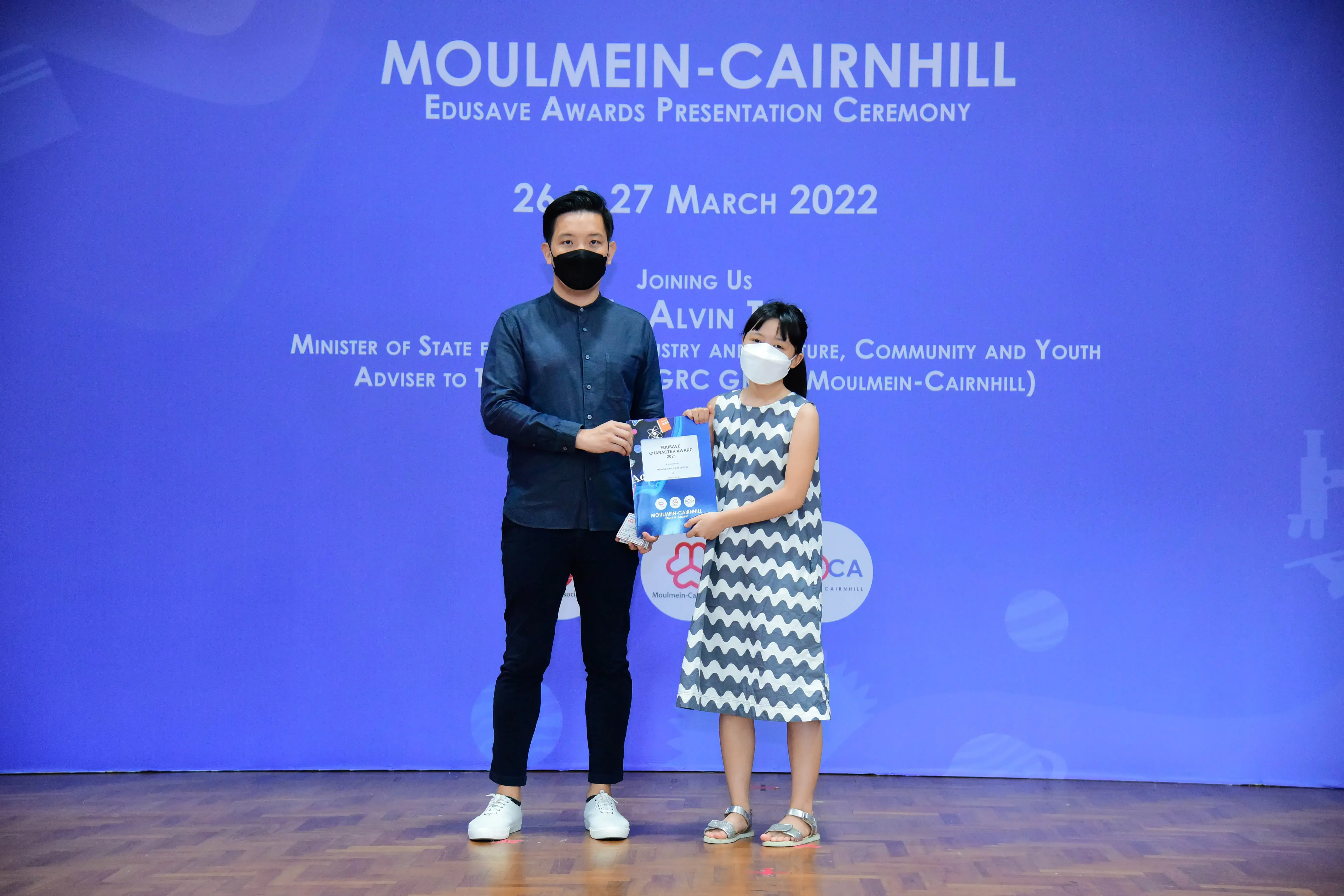 A masked man and a young girl hold a certificate at the Moulmein-Cairnhill Edusave Awards Ceremony, March 2022.