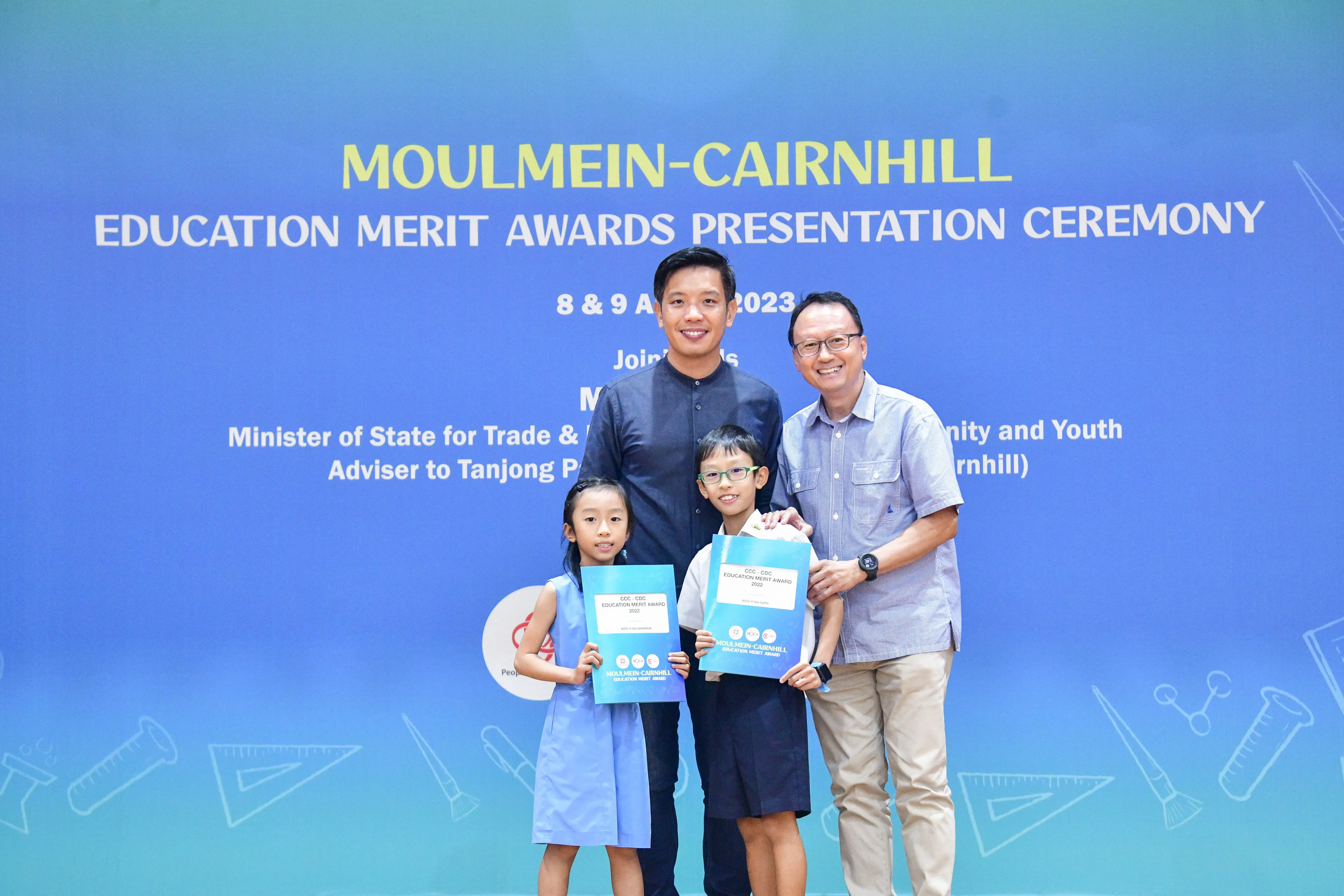 Two men and two children holding award folders pose in front of a Moulmein-Cairnhill Education Merit Awards banner.