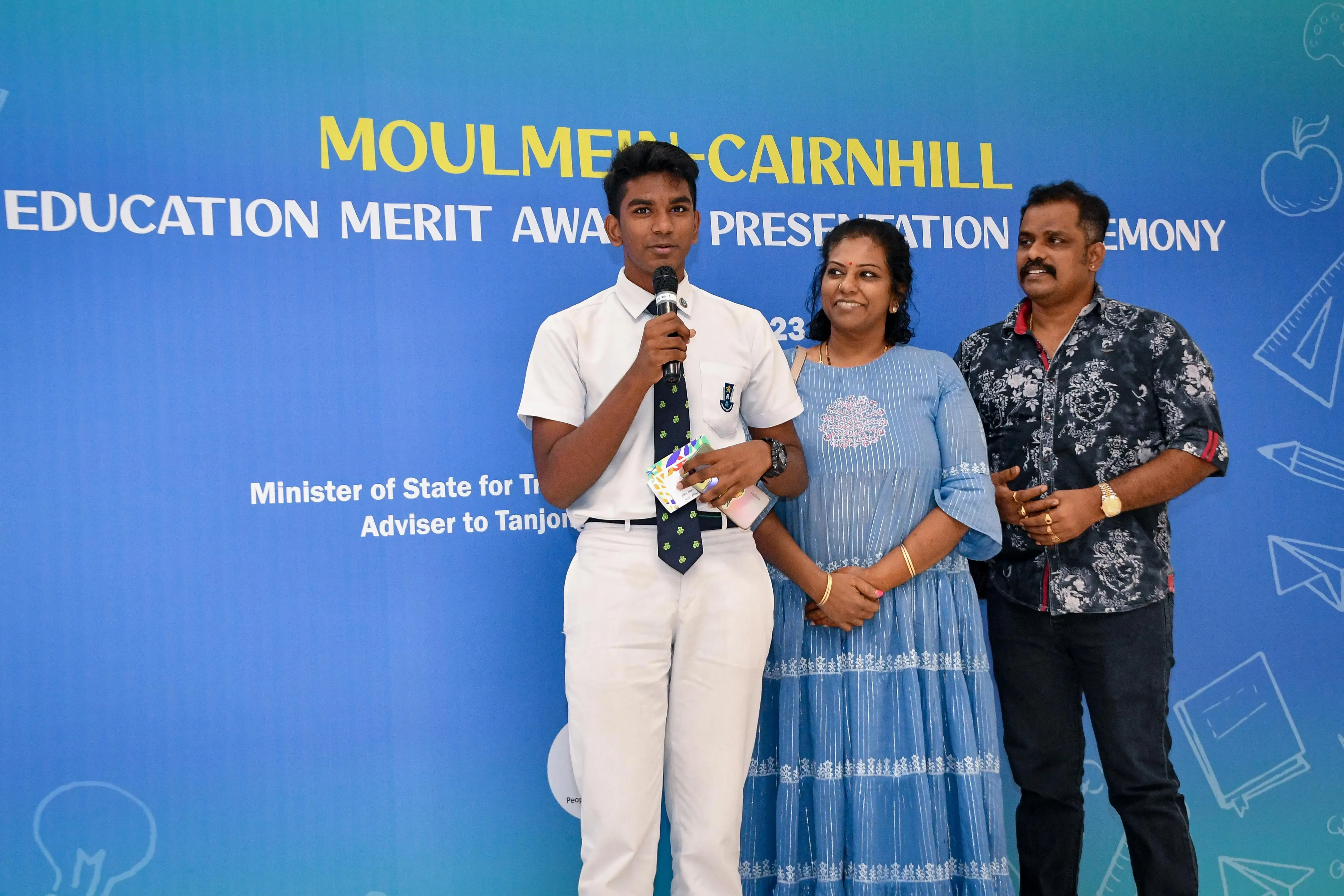 A student in white uniform speaks into a microphone beside two adults at a Moulmein-Cairnhill Education Merit Award ceremony.