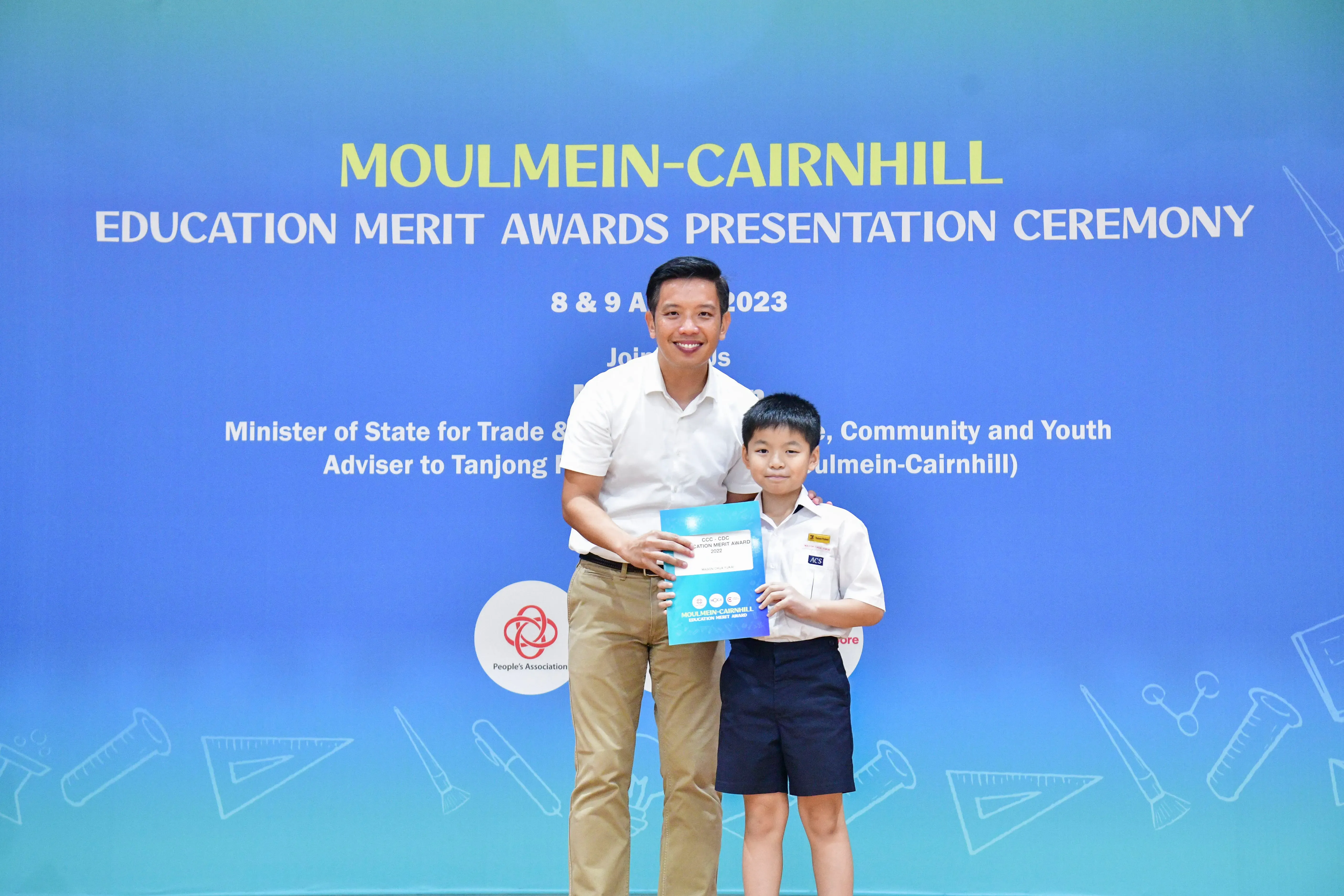 A man and a young boy hold a certificate in front of a Moulmein-Cairnhill Education Merit Awards banner.
