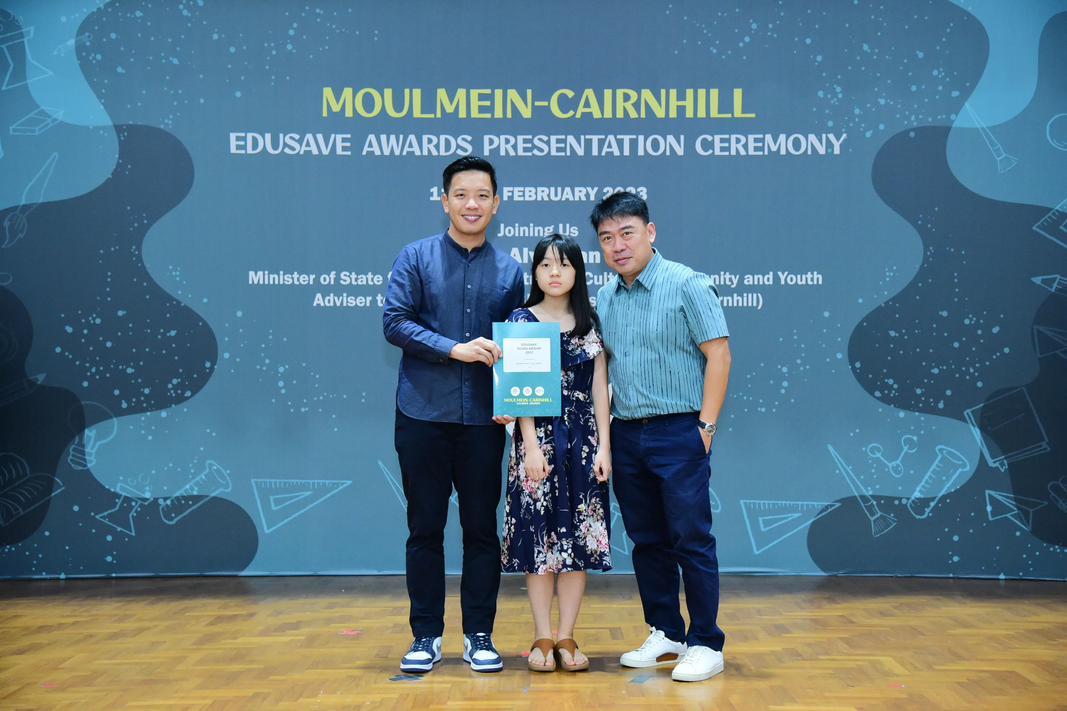 Two men and a girl holding a certificate at the Moulmein-Cairnhill Edusave Awards Presentation Ceremony backdrop.