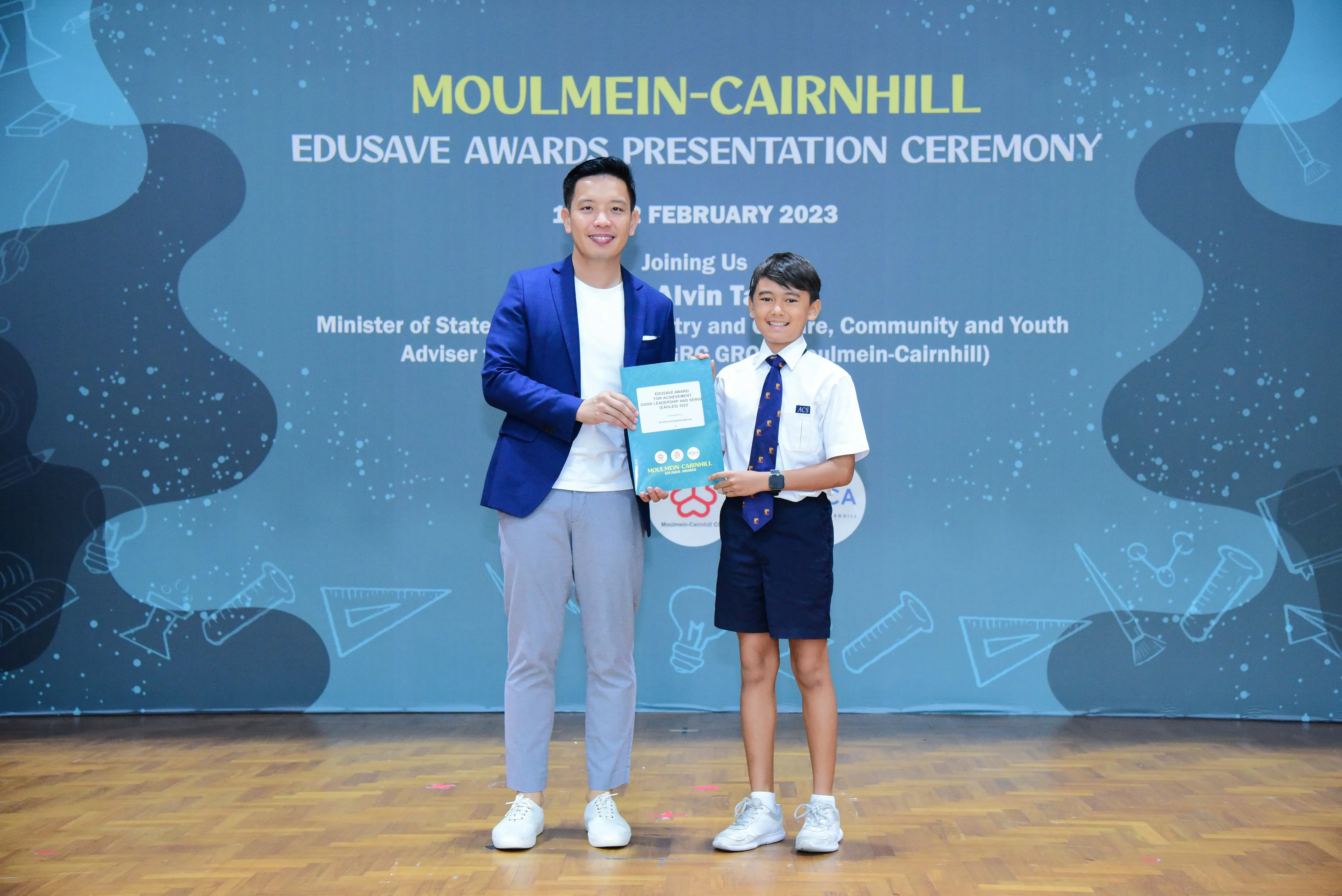 An adult in a blue blazer and a uniformed boy hold an award together at the Moulmein-Cairnhill Edusave Awards Ceremony 2023.