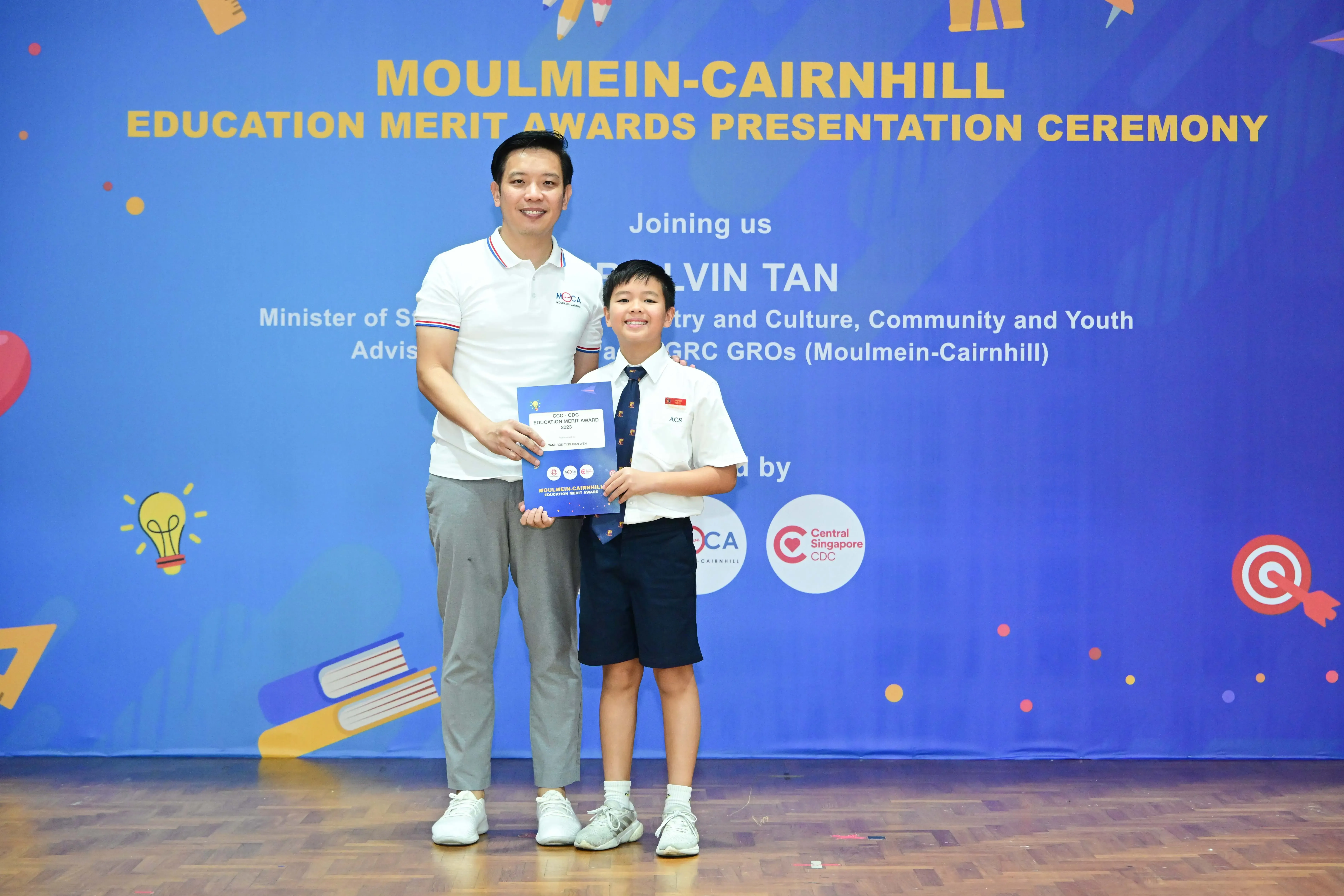 A man and a young boy in school uniform hold a certificate at the Moulmein-Cairnhill Education Merit Awards ceremony.
