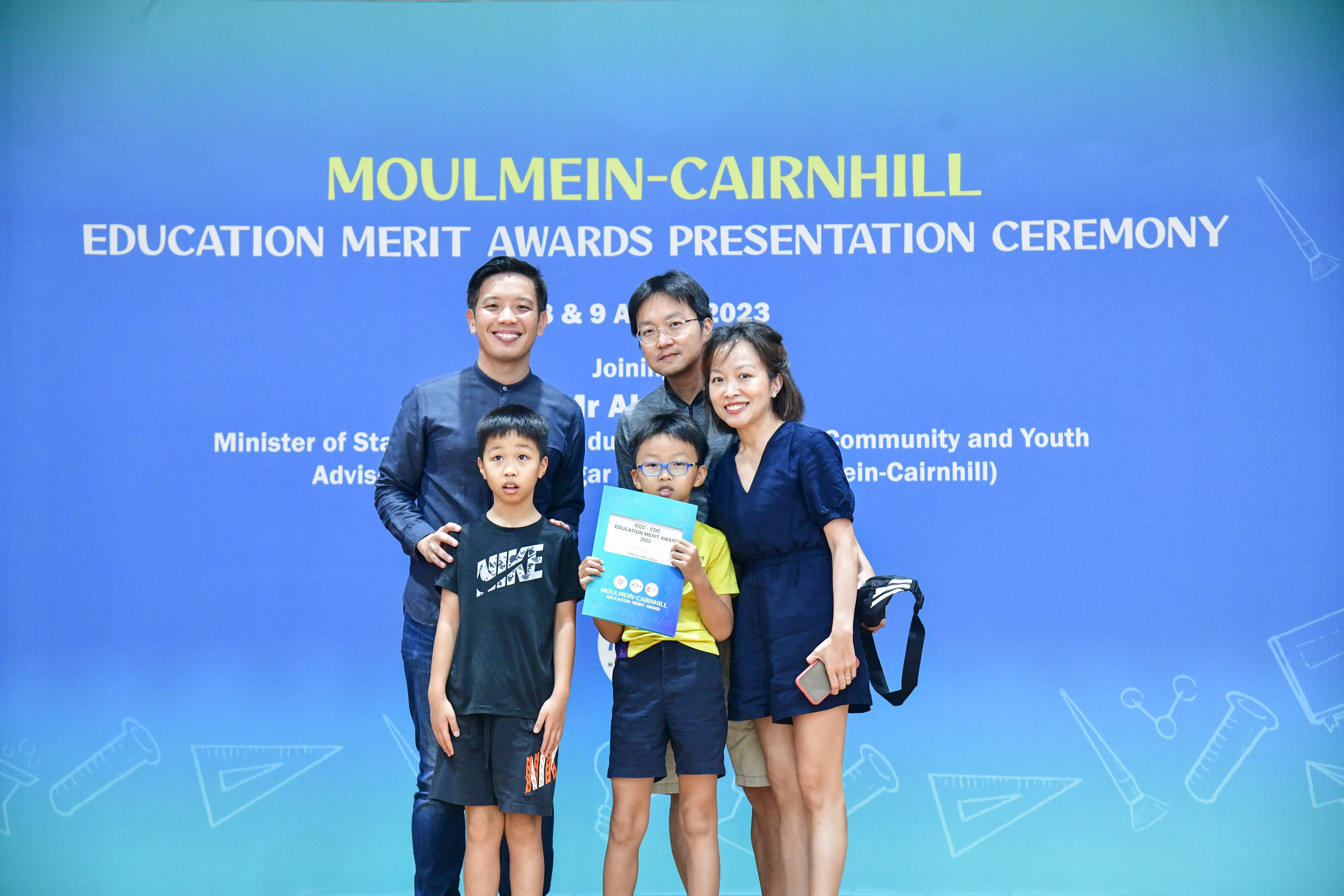 Four adults and two children pose before a Moulmein-Cairnhill Education Merit Awards Ceremony banner in 2023.