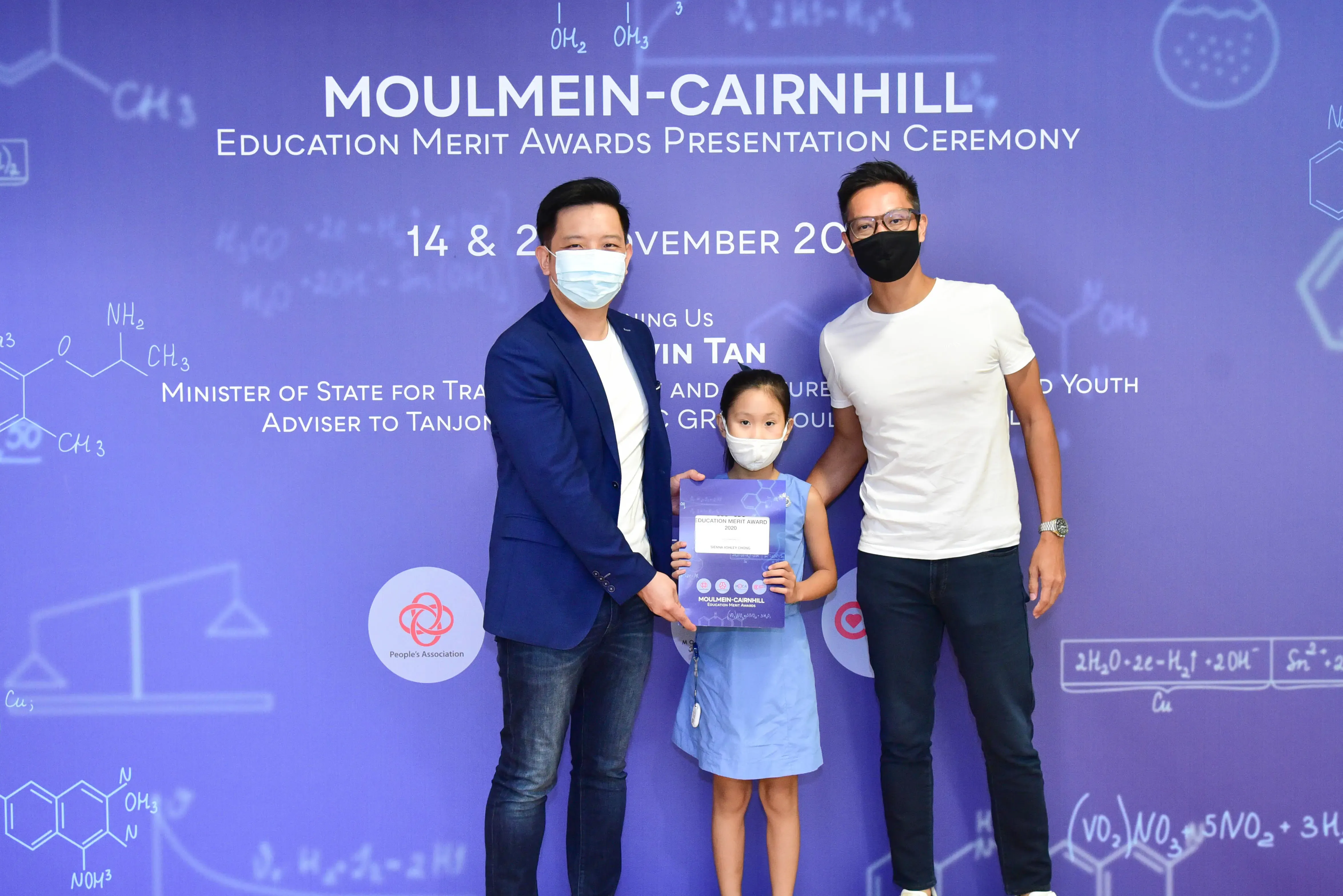 Two masked men and a young girl holding an award certificate pose at the Moulmein-Cairnhill Education Merit Awards ceremony.