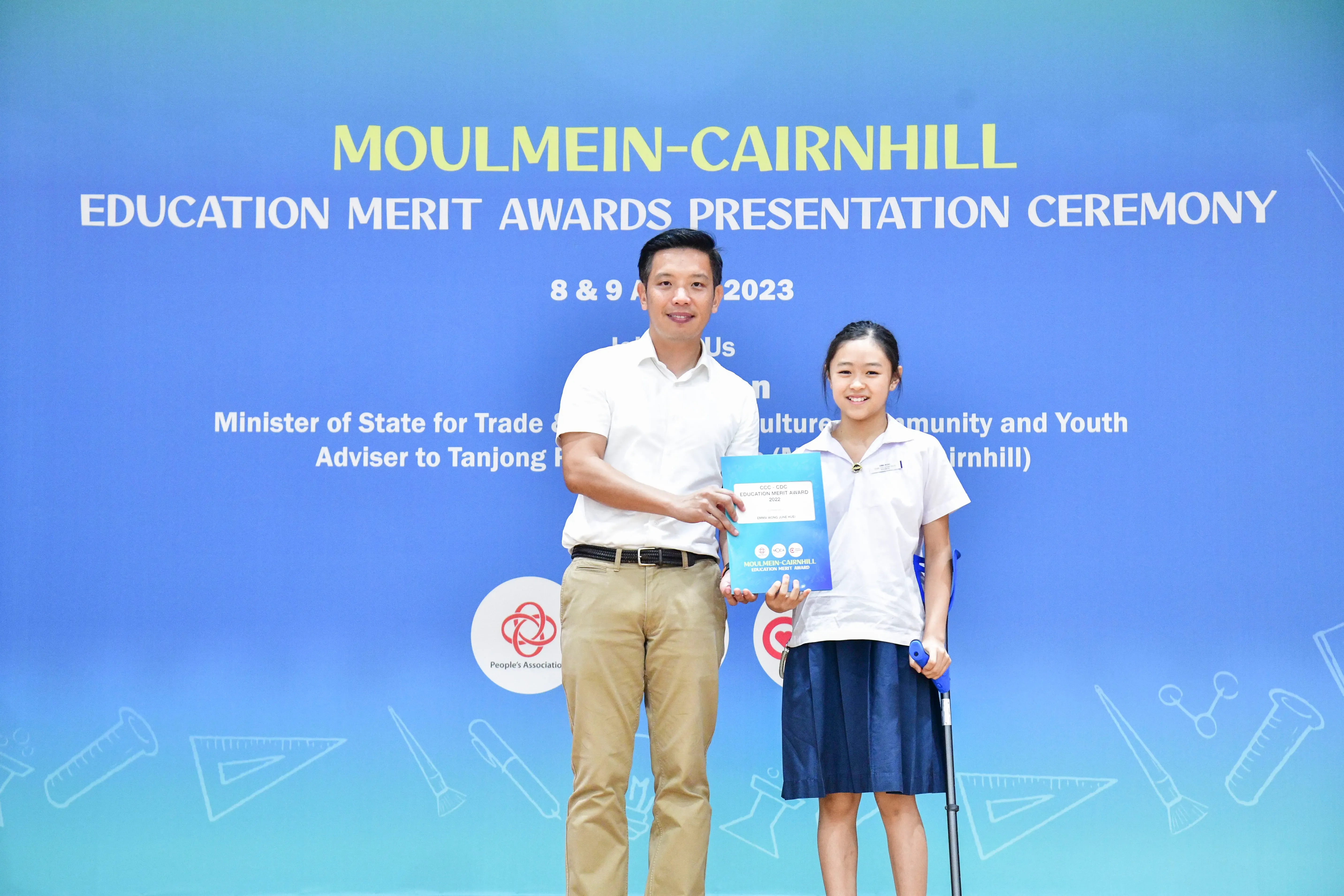 Man and young girl holding a certificate in front of a Moulmein-Cairnhill Education Merit Awards 2023 banner.