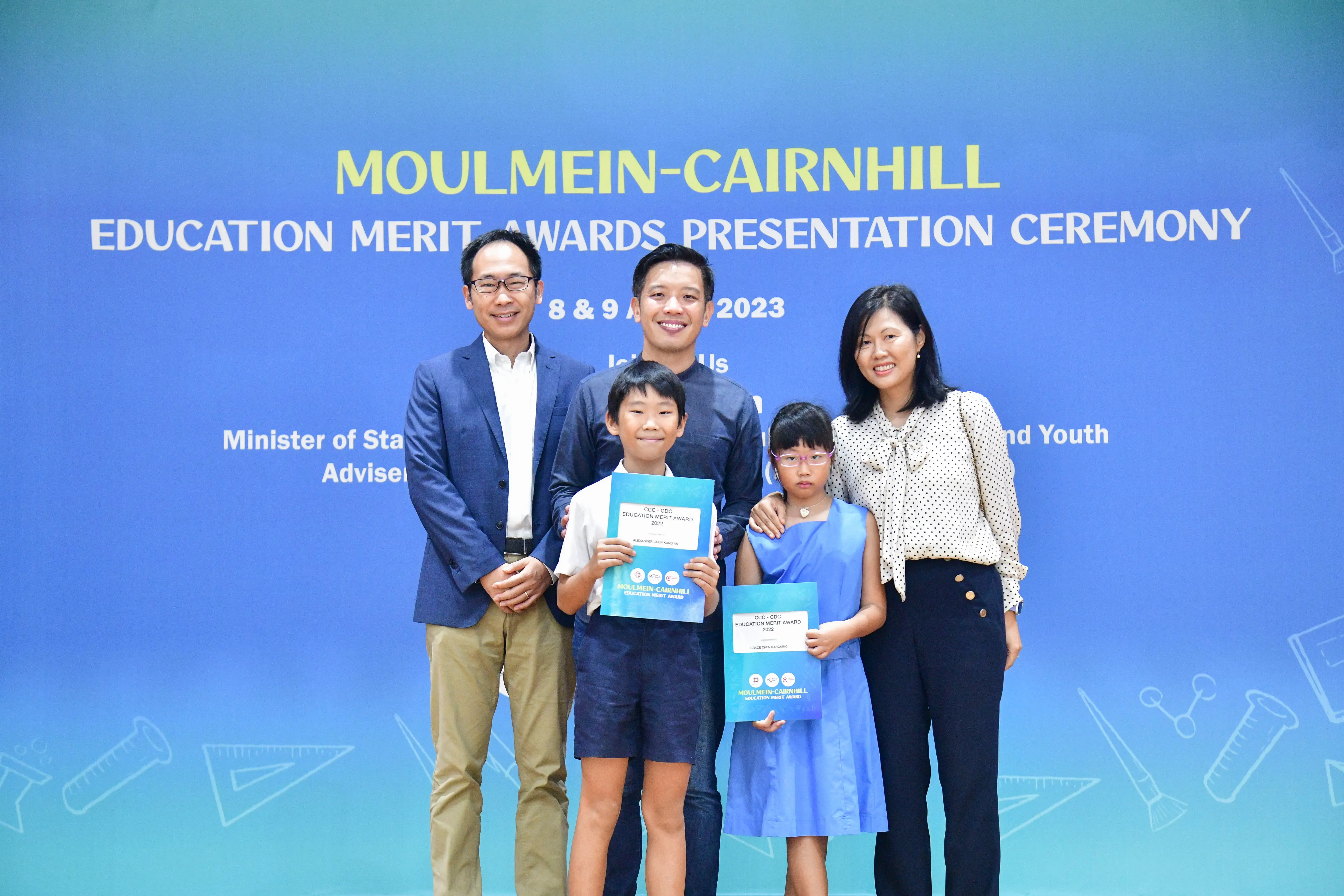 Five people pose before a Moulmein-Cairnhill Education Merit Awards 2023 banner, two children holding blue award certificates.