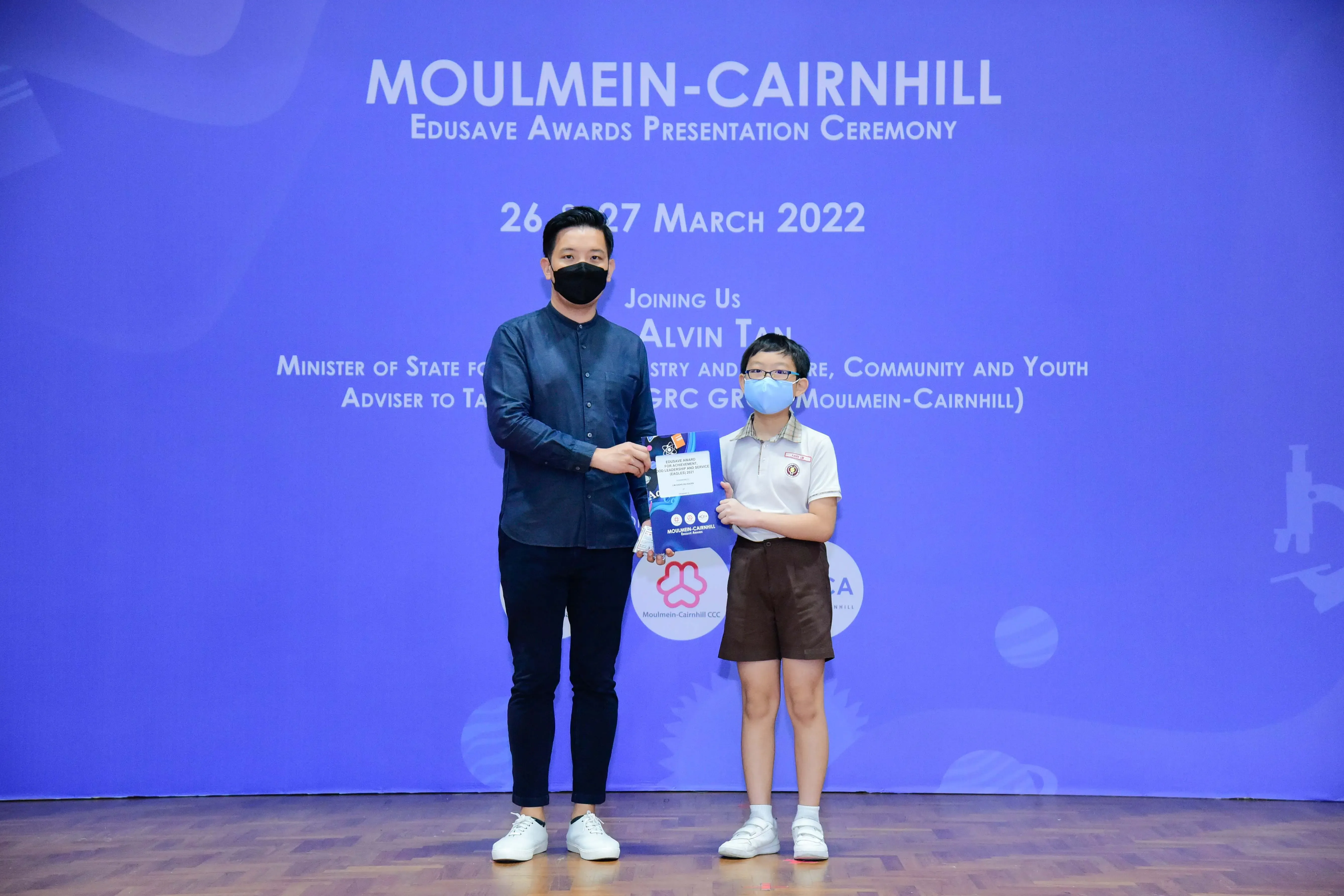 An adult and a young student in masks hold an award at the Moulmein-Cairnhill Edusave Awards Ceremony, March 2022.