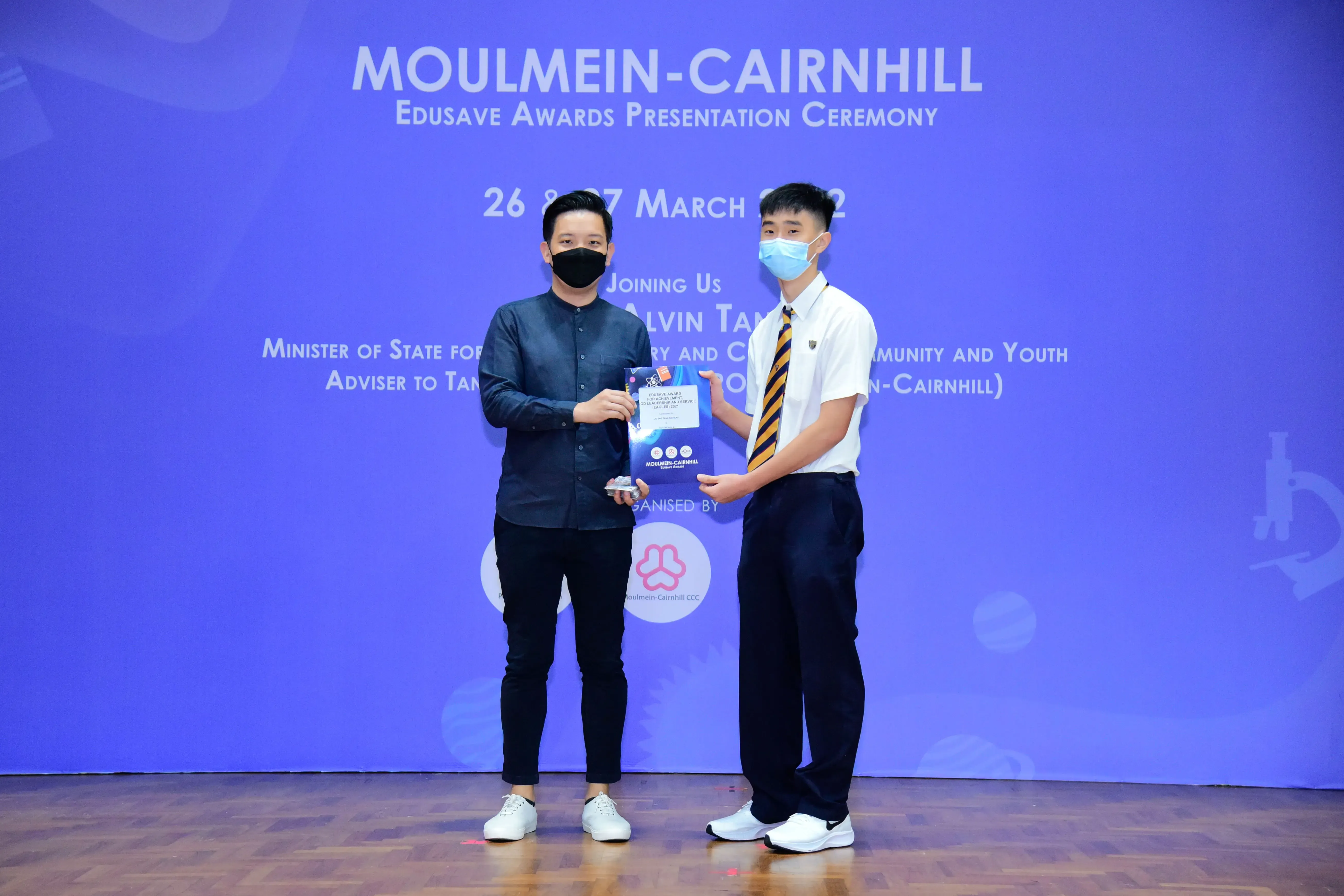 Two masked men hold a certificate at the Moulmein-Cairnhill Edusave Awards Presentation Ceremony backdrop.