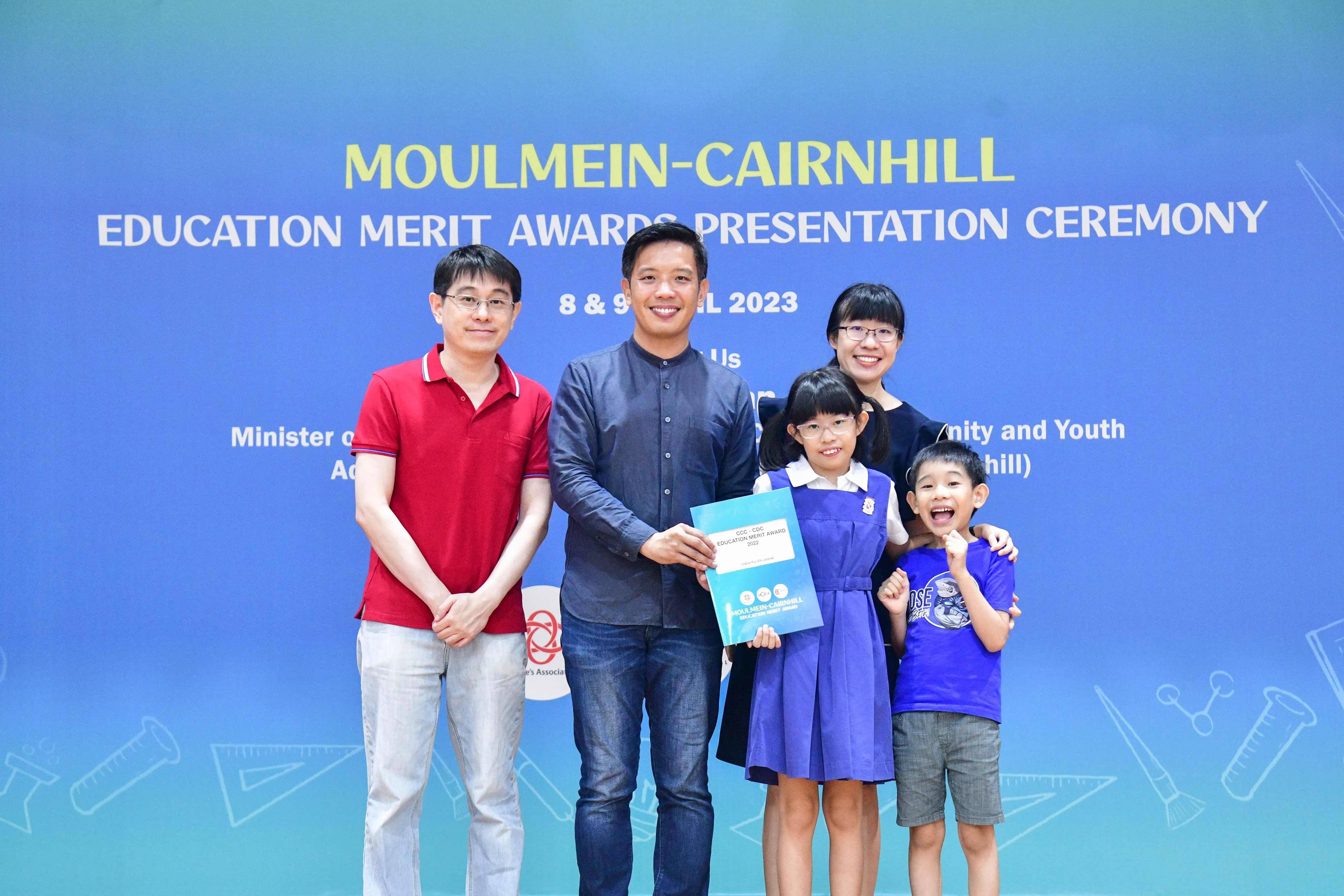 Five people pose before a Moulmein-Cairnhill Education Merit Awards 2023 banner, with a girl holding a certificate.