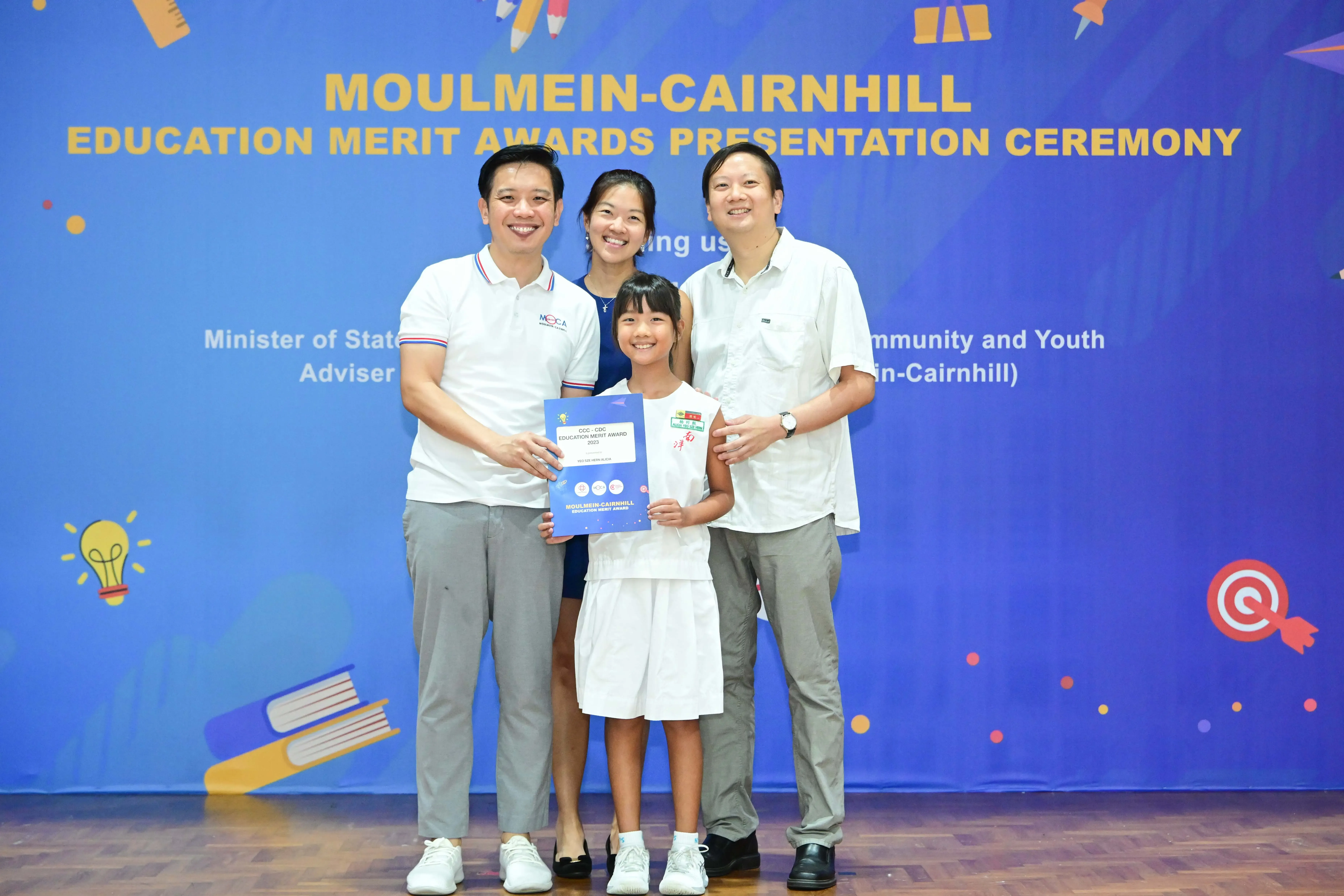 Four people pose smiling before a blue Moulmein-Cairnhill Education Merit Awards banner, with a girl holding a certificate.