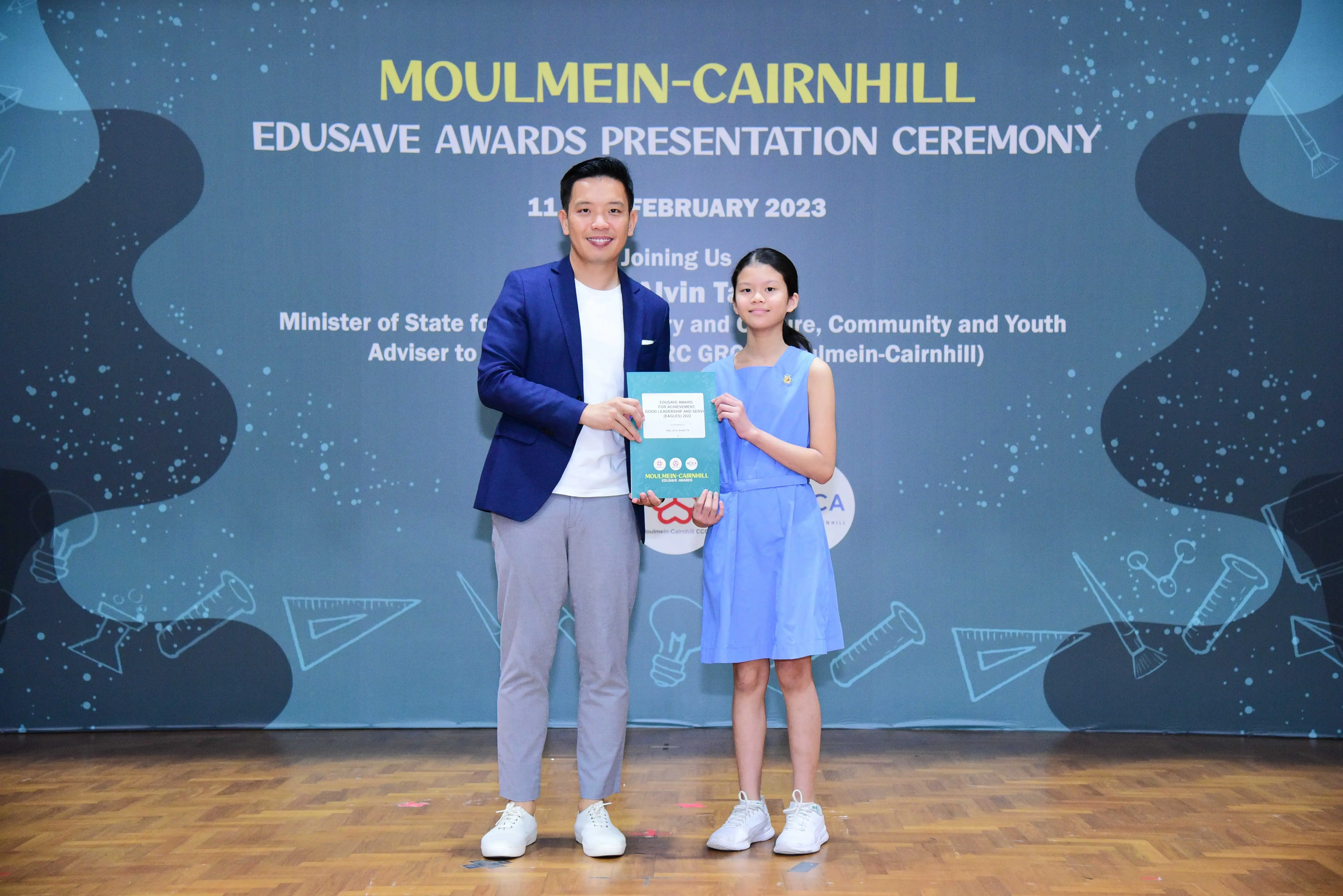 A man in a blue blazer and a young girl in a blue dress hold a certificate at the Moulmein-Cairnhill Edusave Awards 2023.