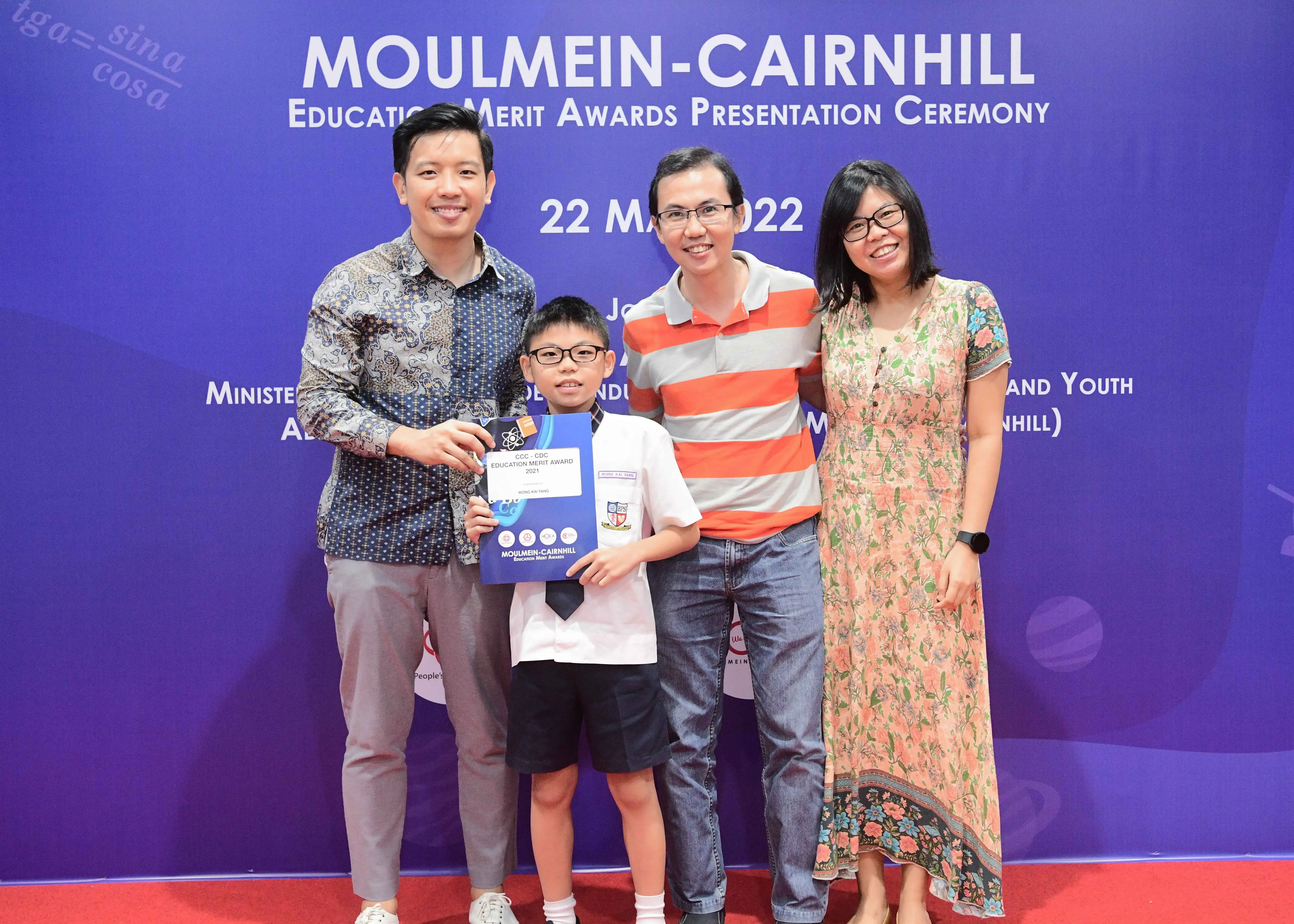 Four people pose smiling before a Moulmein-Cairnhill Education Merit Awards banner; a boy holds an award certificate.