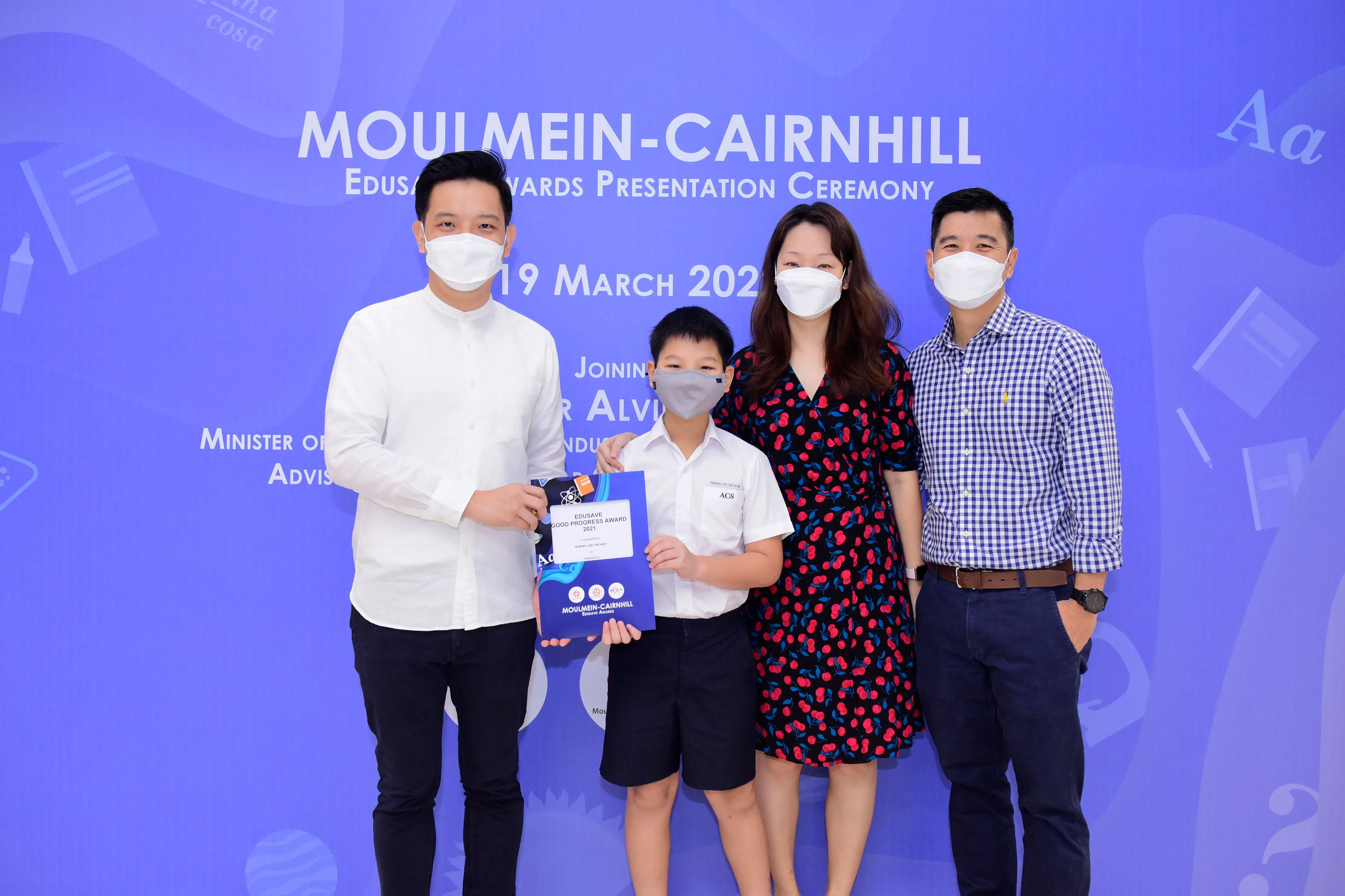 Four masked people pose at the Moulmein-Cairnhill Edusave Awards Presentation Ceremony, with a boy holding a certificate.