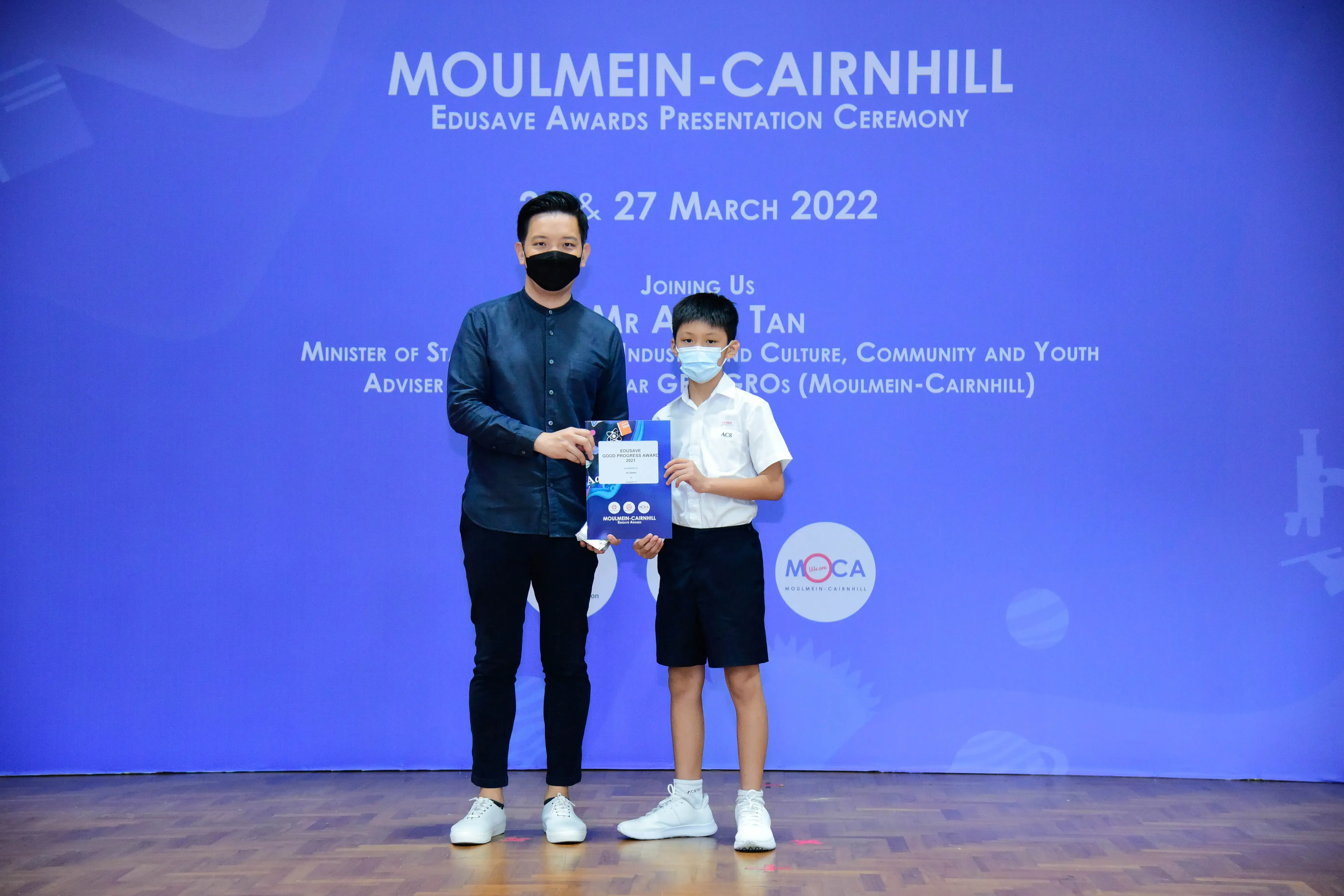 An adult and a young boy wearing masks hold a certificate at the Moulmein-Cairnhill Edusave Awards Ceremony 2022.