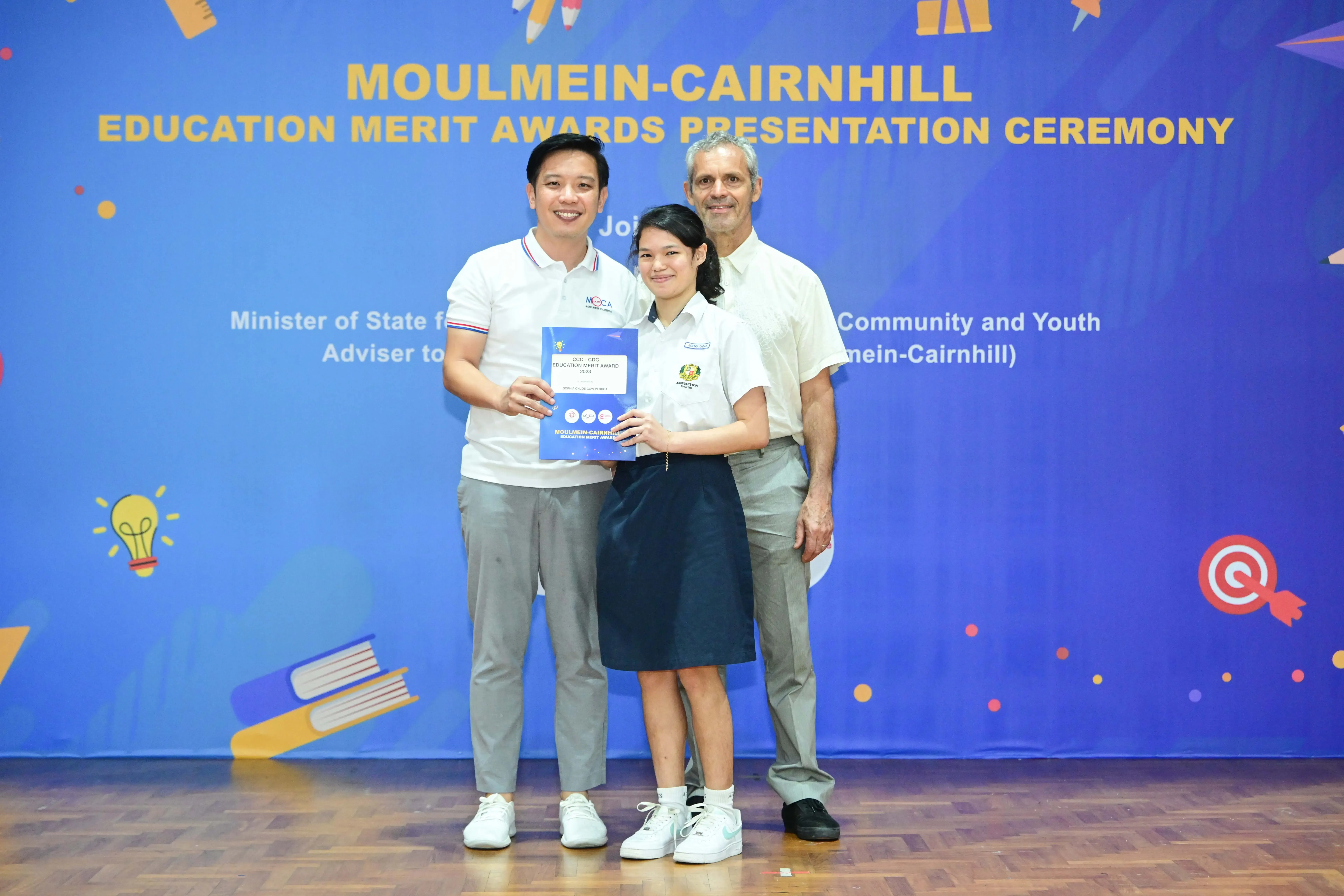 Three people pose holding a certificate in front of a blue Moulmein-Cairnhill Education Merit Awards banner.