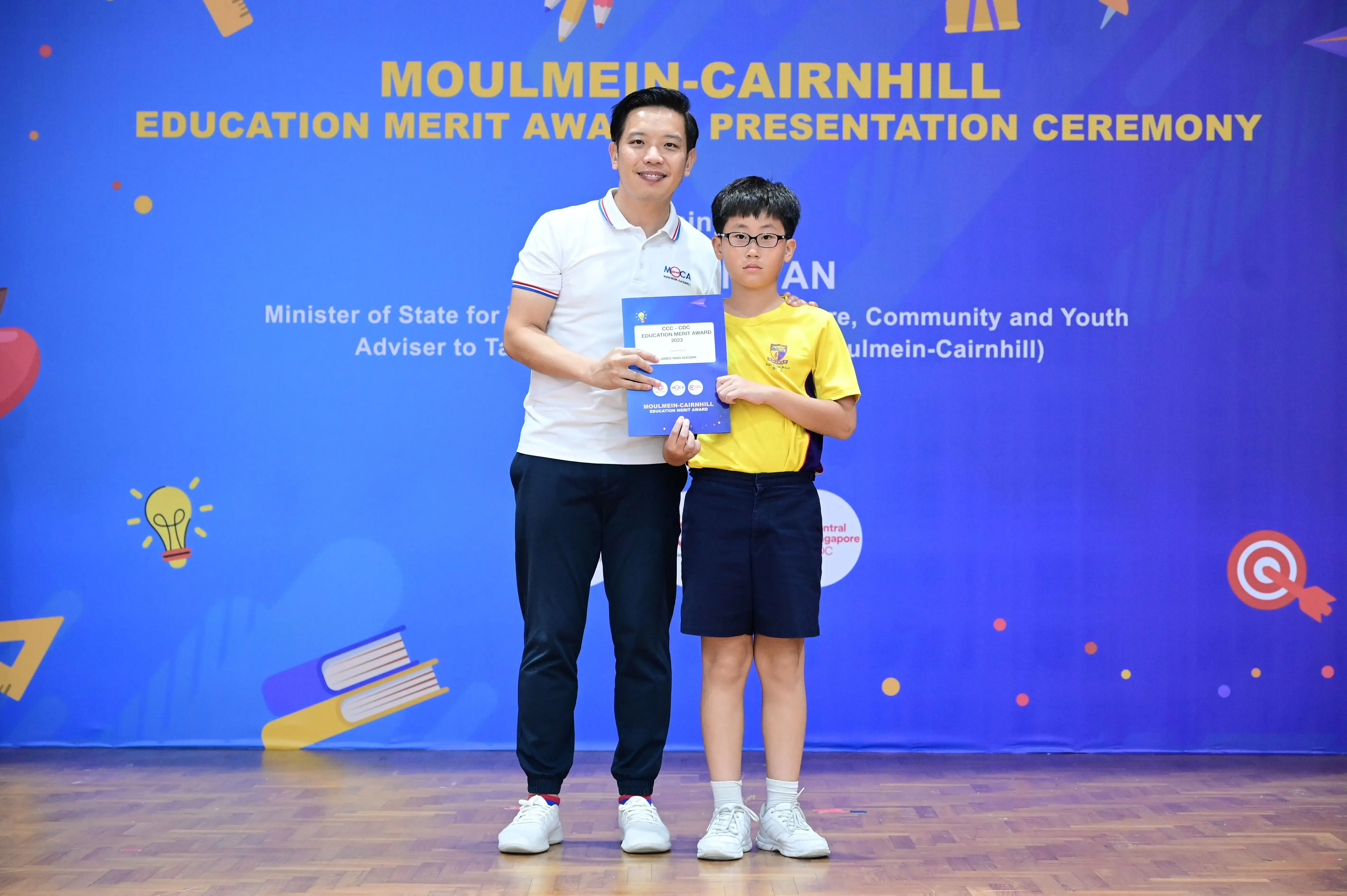 An adult and a young boy hold an award certificate at the Moulmein-Cairnhill Education Merit Award Presentation Ceremony.