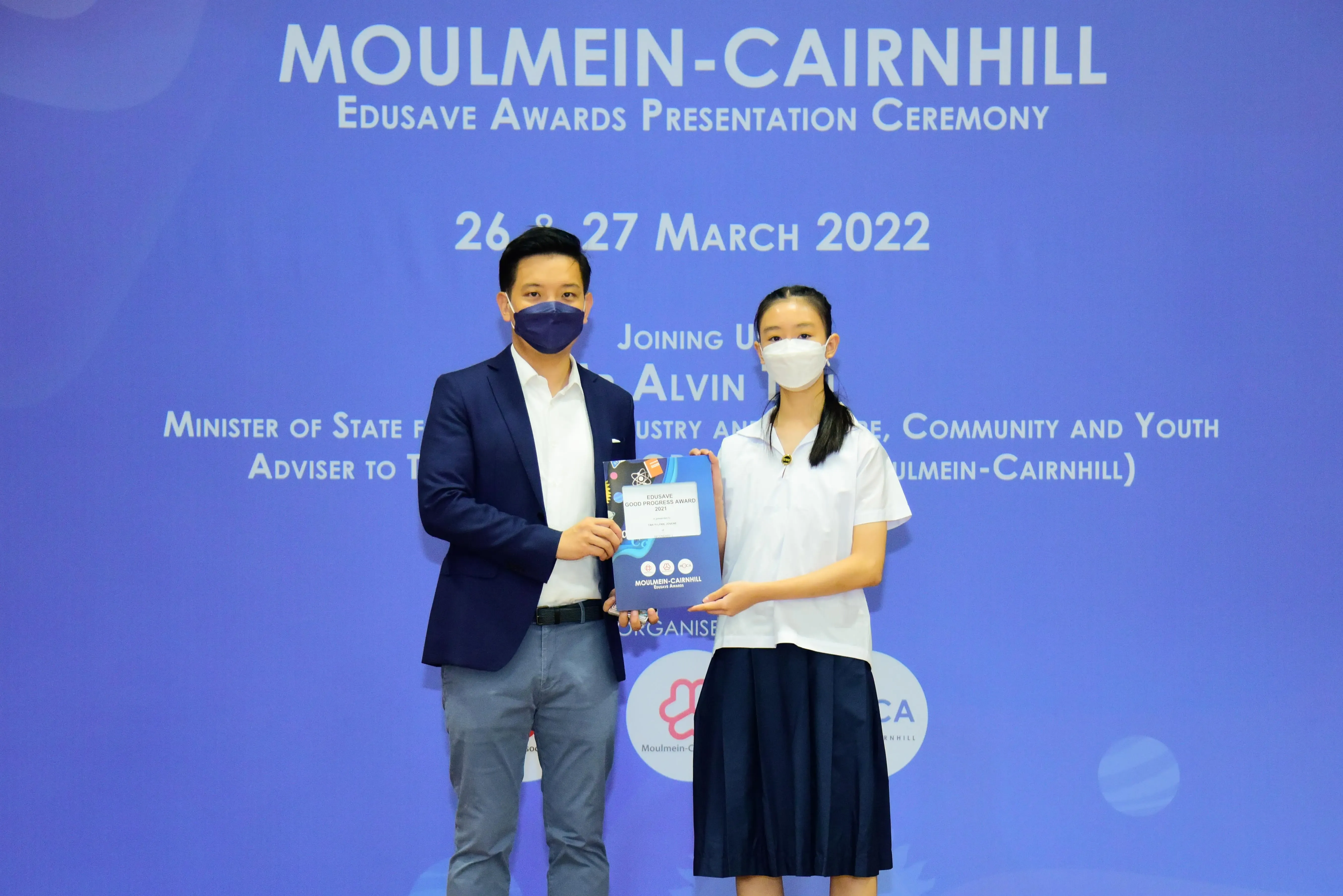 Two masked individuals hold an award folder at the Moulmein-Cairnhill Edusave Awards Presentation Ceremony, March 2022.