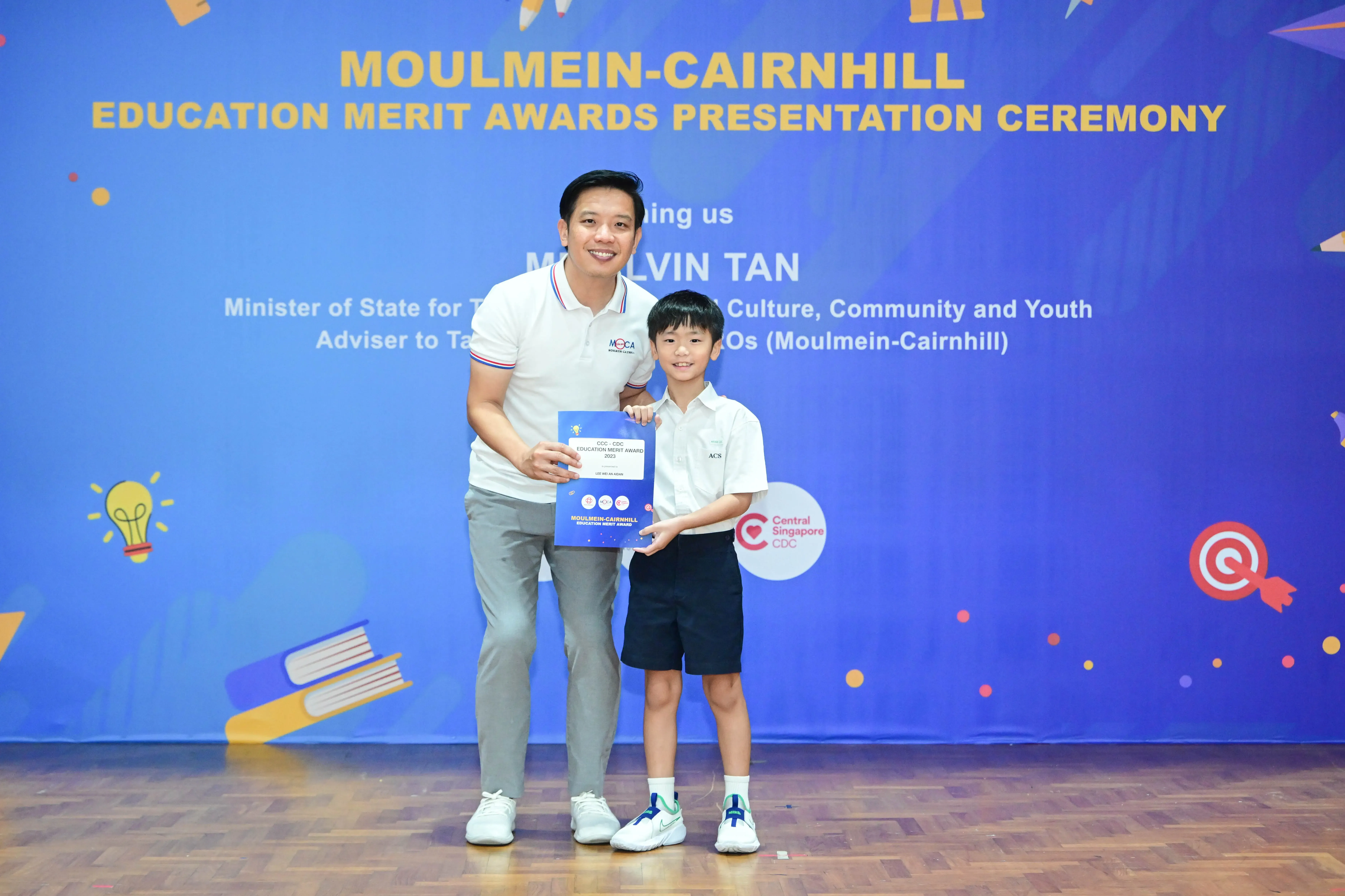 Man and young boy hold a blue award certificate at Moulmein-Cairnhill Education Merit Awards Presentation Ceremony.