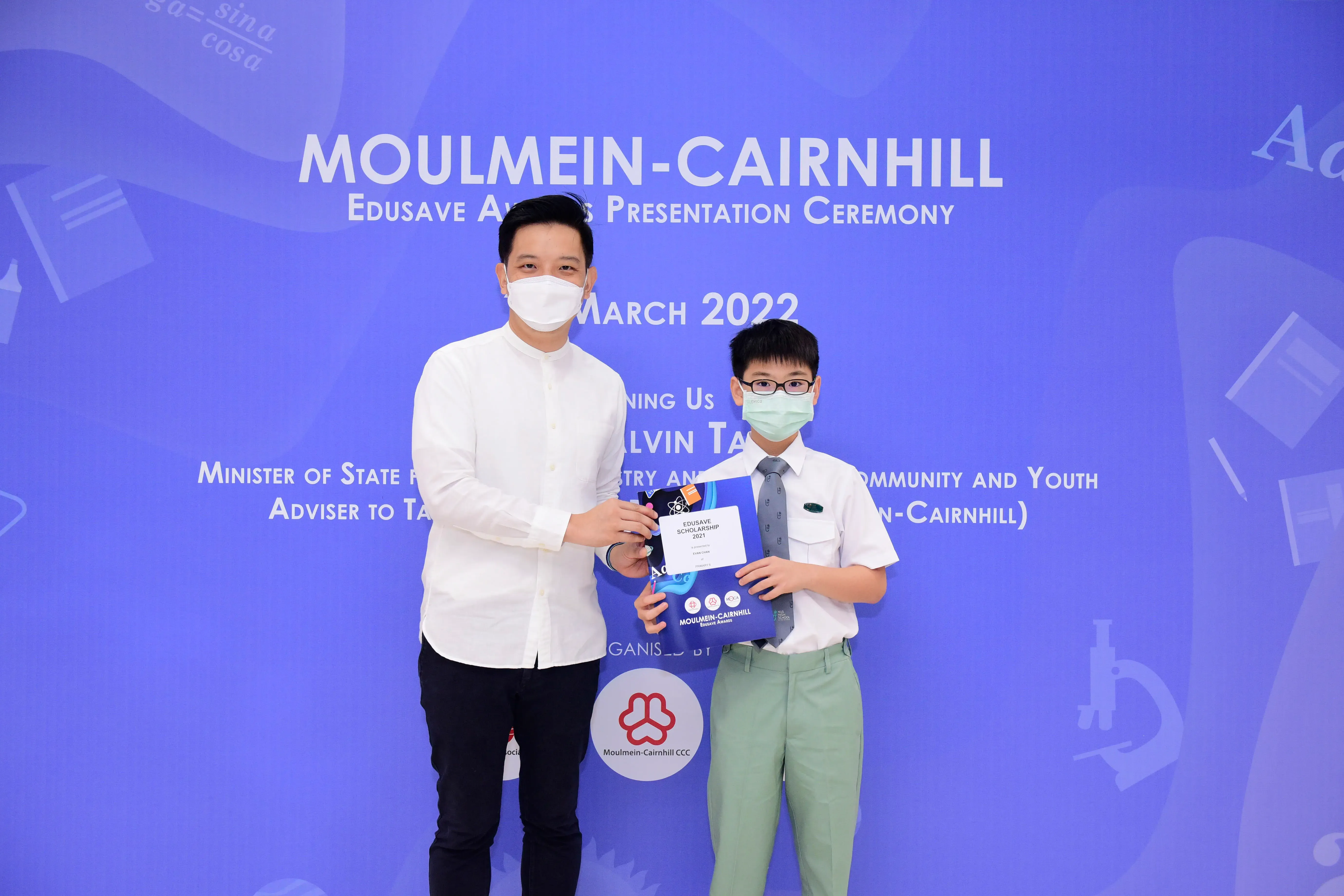 An adult and a masked schoolboy hold an Edusave Scholarship certificate at the Moulmein-Cairnhill ceremony, March 2022.
