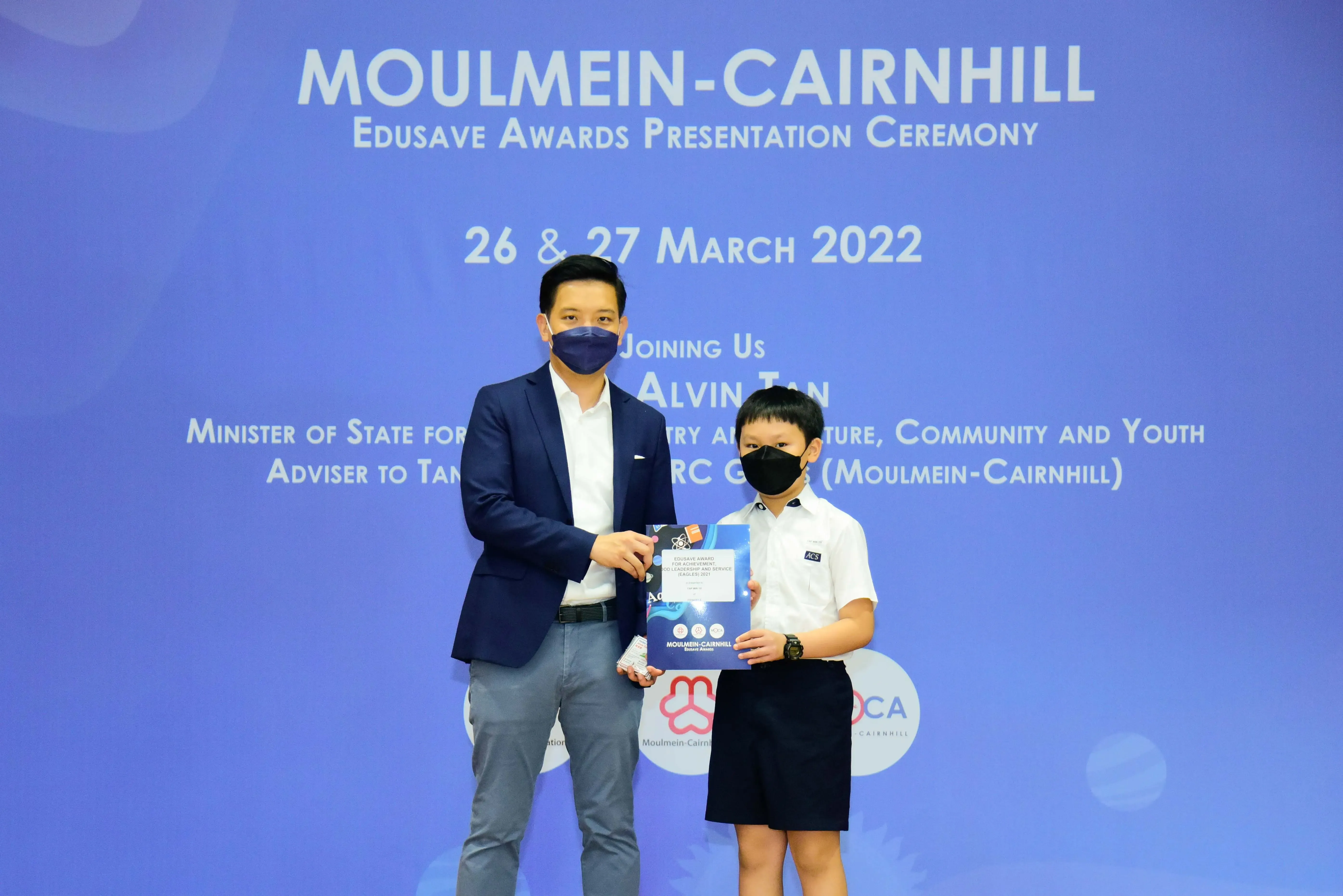 An adult and a young student in masks hold an award certificate at the Moulmein-Cairnhill Edusave Awards Ceremony 2022.