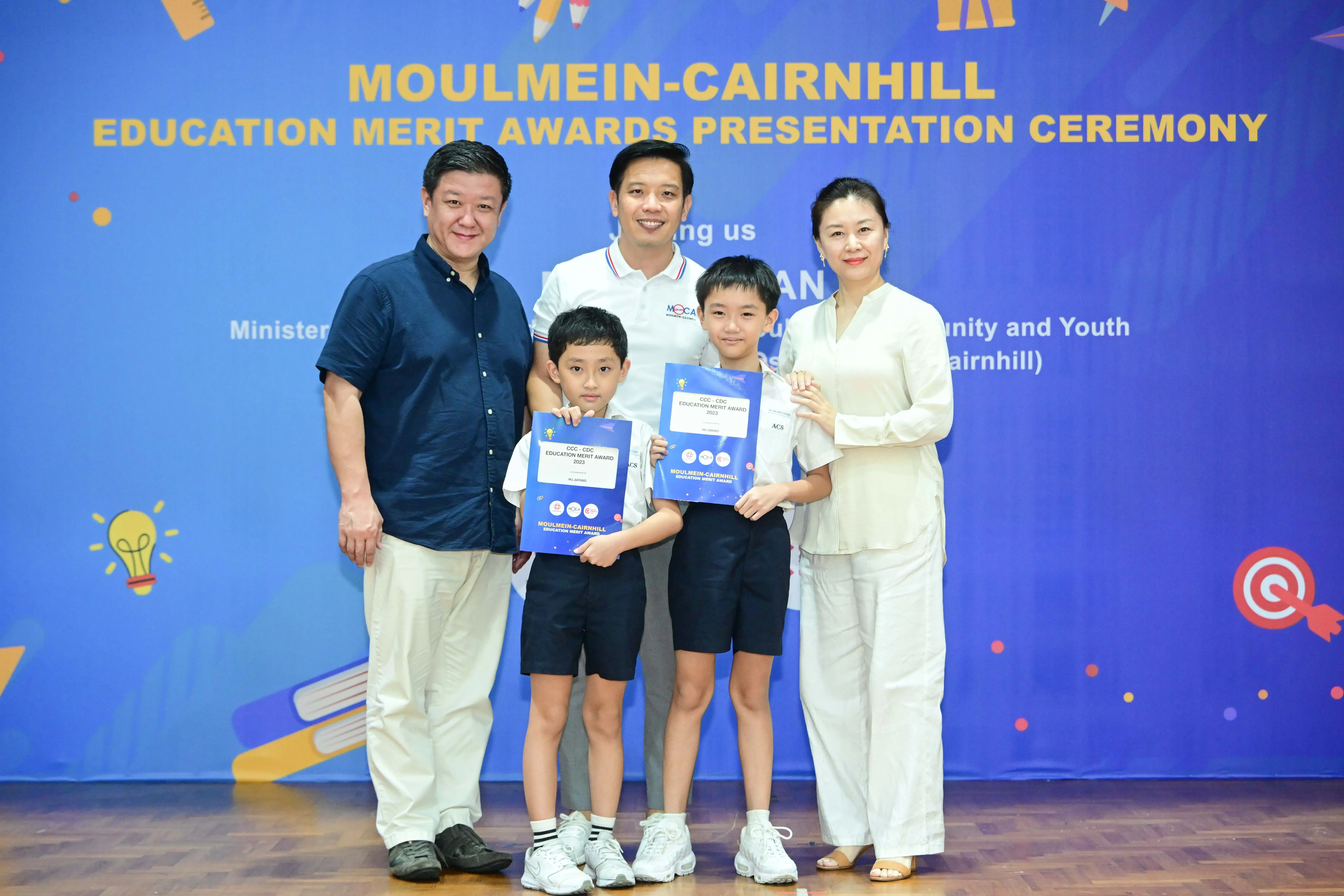 Two boys holding award certificates pose with three adults at the Moulmein-Cairnhill Education Merit Awards Ceremony.