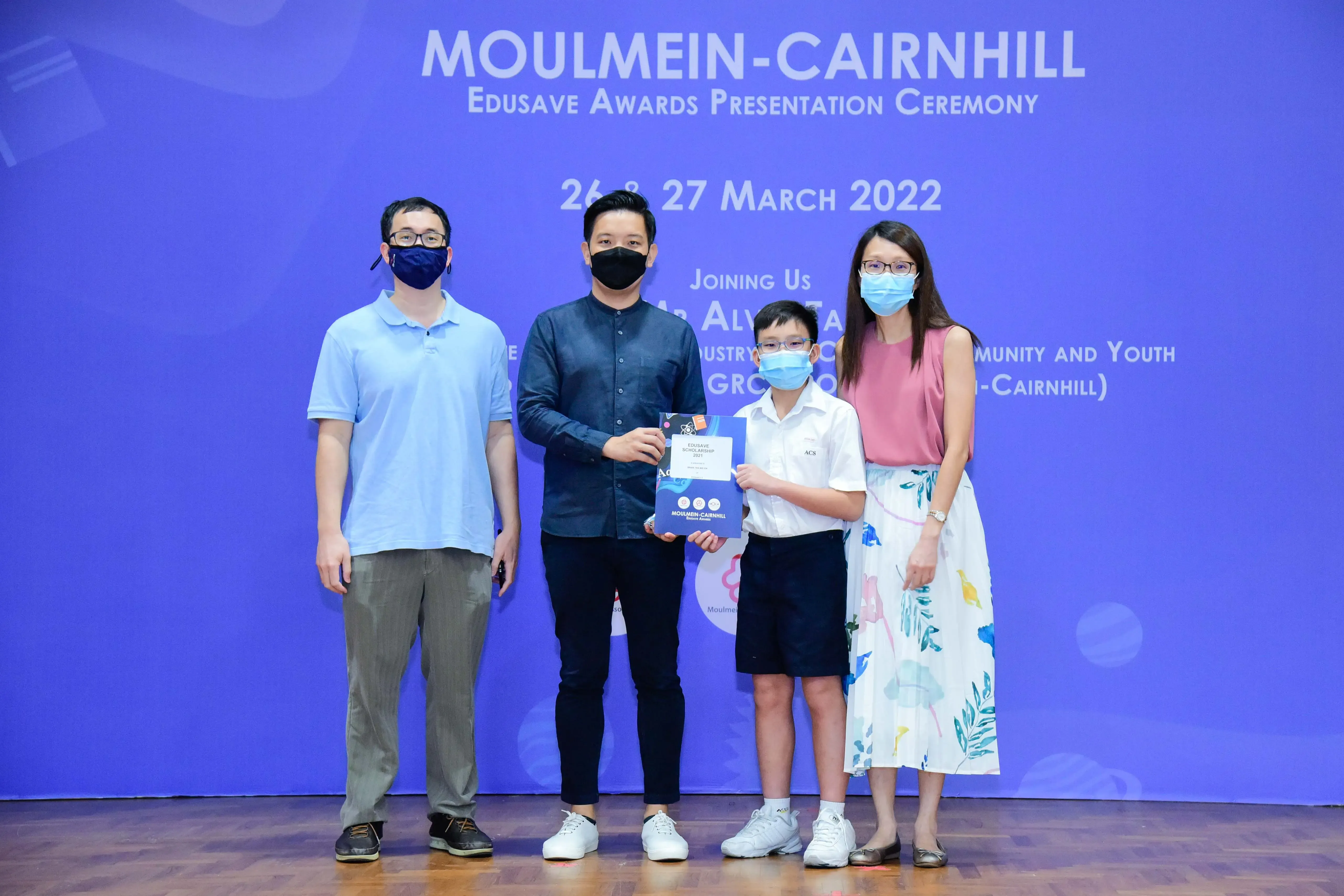 Four masked people stand before a blue Moulmein-Cairnhill Edusave Awards Presentation Ceremony banner, holding a certificate.
