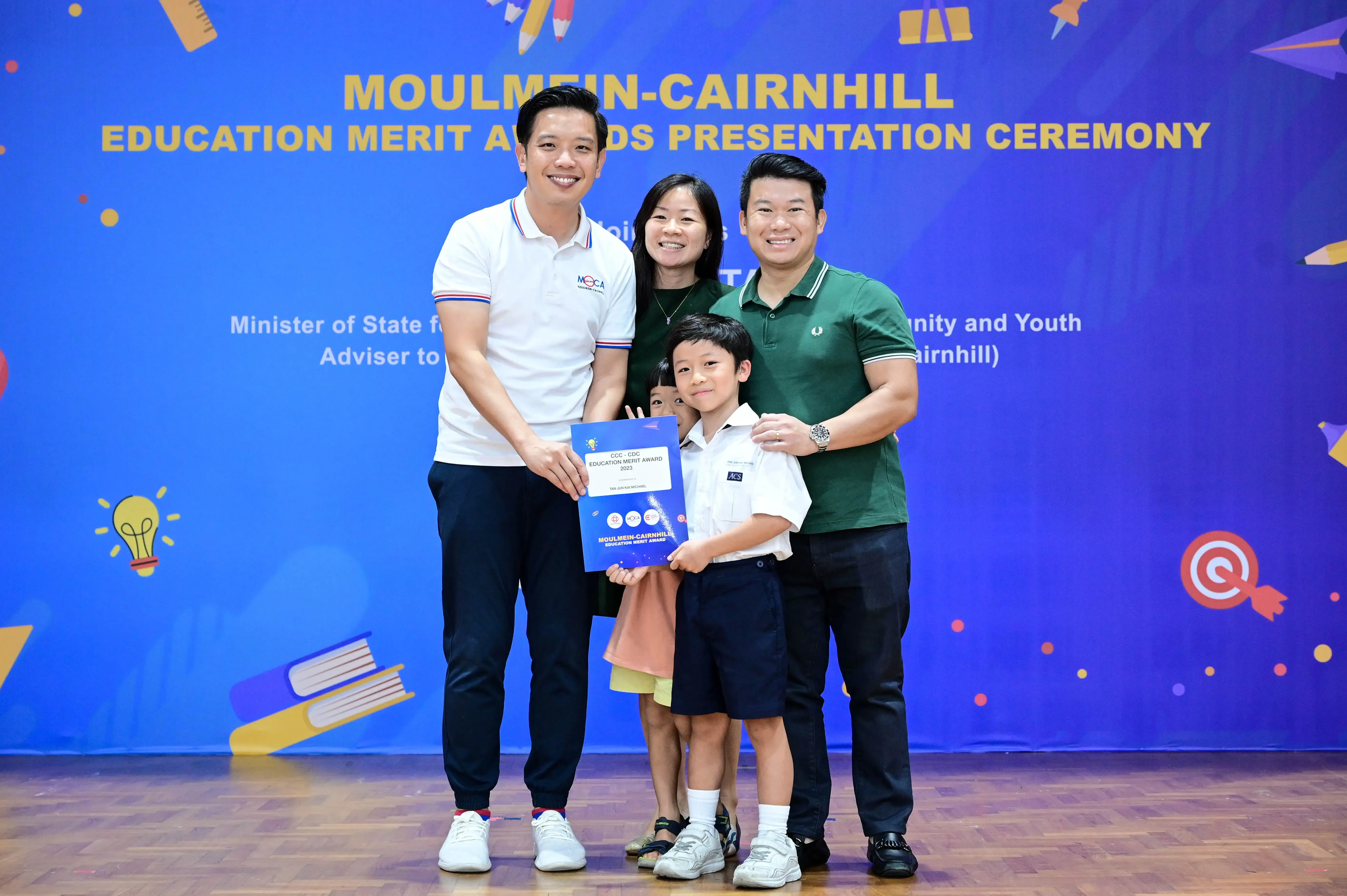 A boy holds an award certificate with two men and a woman smiling at the Moulmein-Cairnhill Education Merit Awards ceremony.
