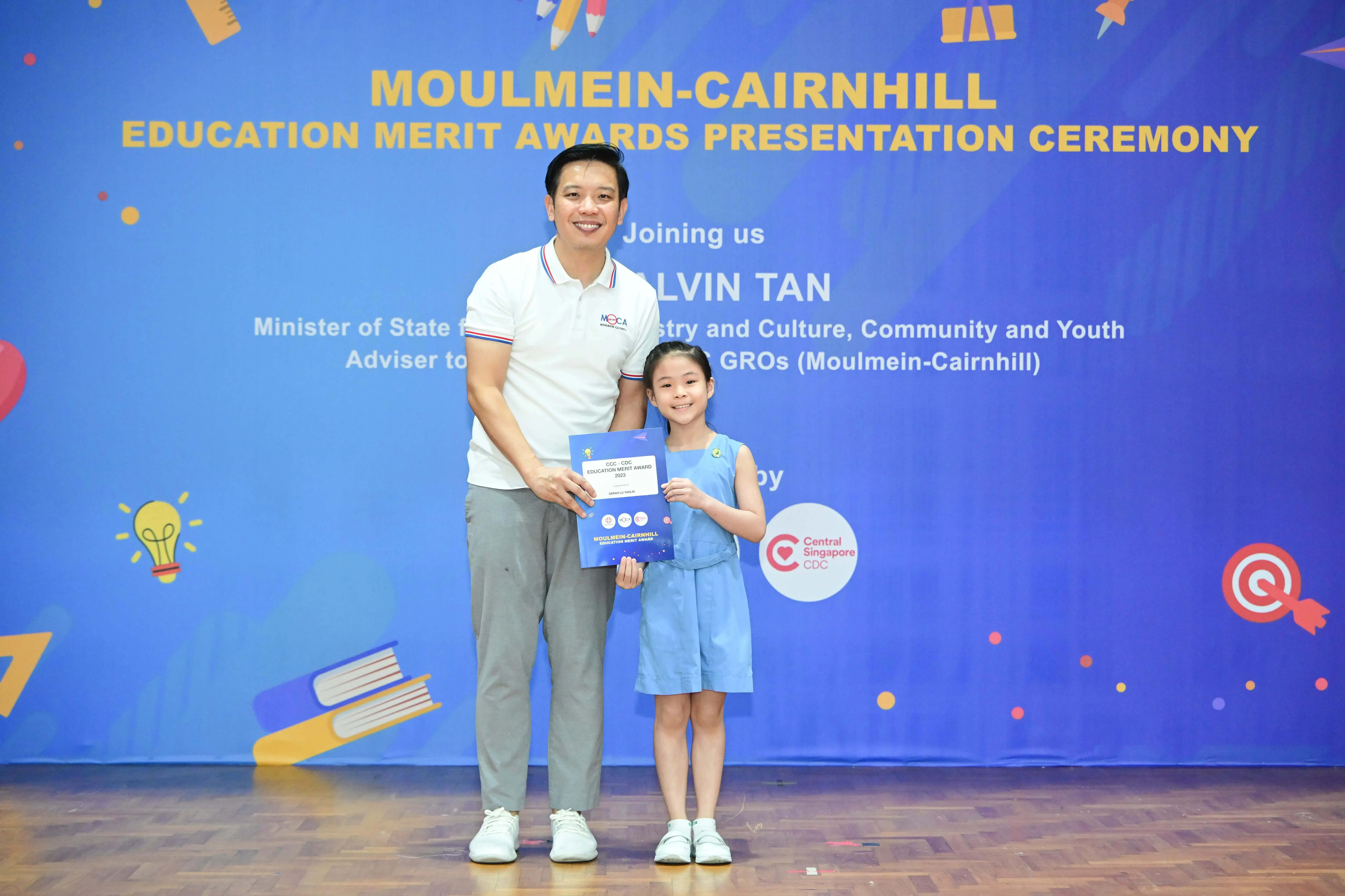 Man and young girl holding a blue award certificate in front of a Moulmein-Cairnhill Education Merit Awards banner.