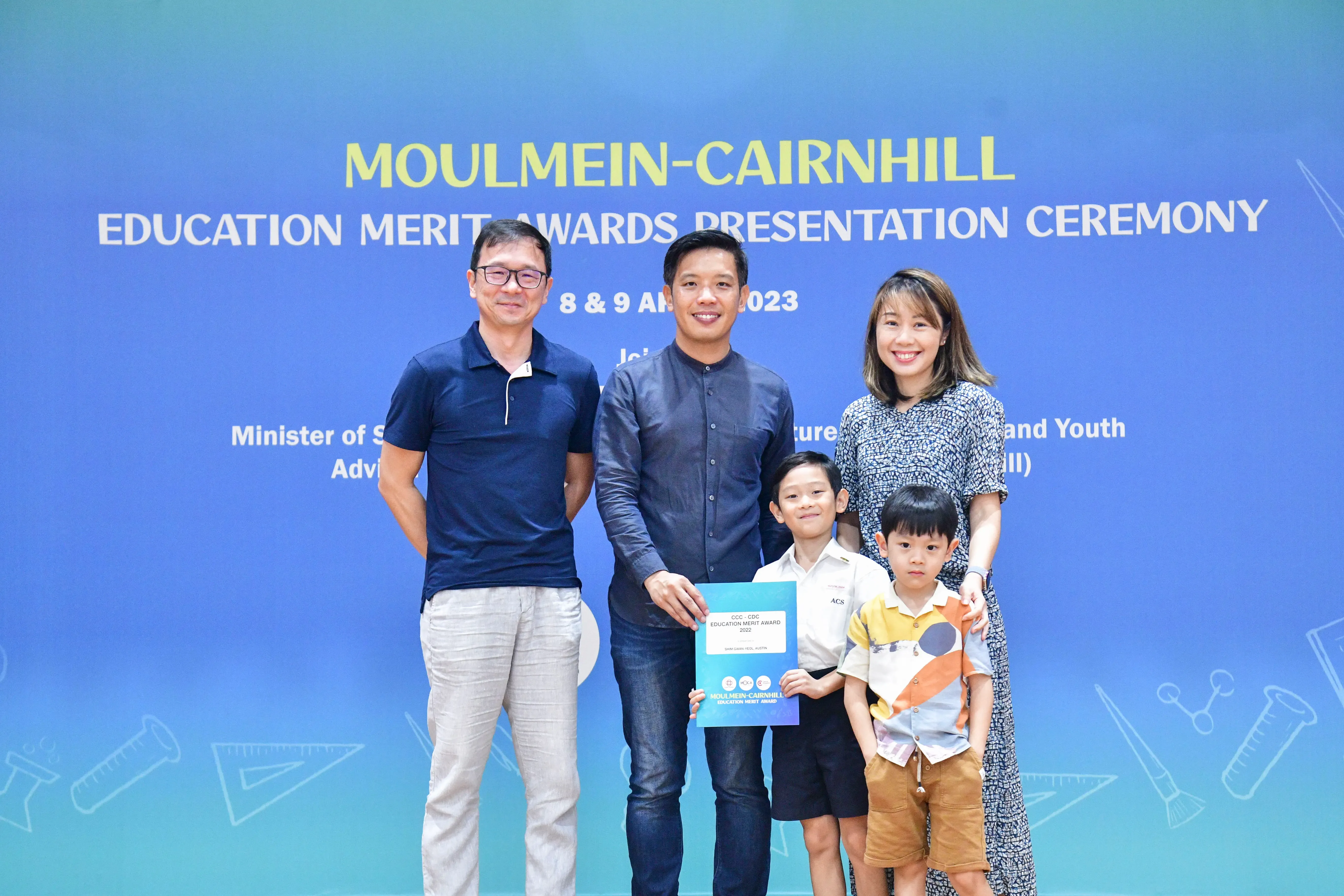Three adults and two young boys pose holding an award certificate at the Moulmein-Cairnhill Education Merit Awards 2023.