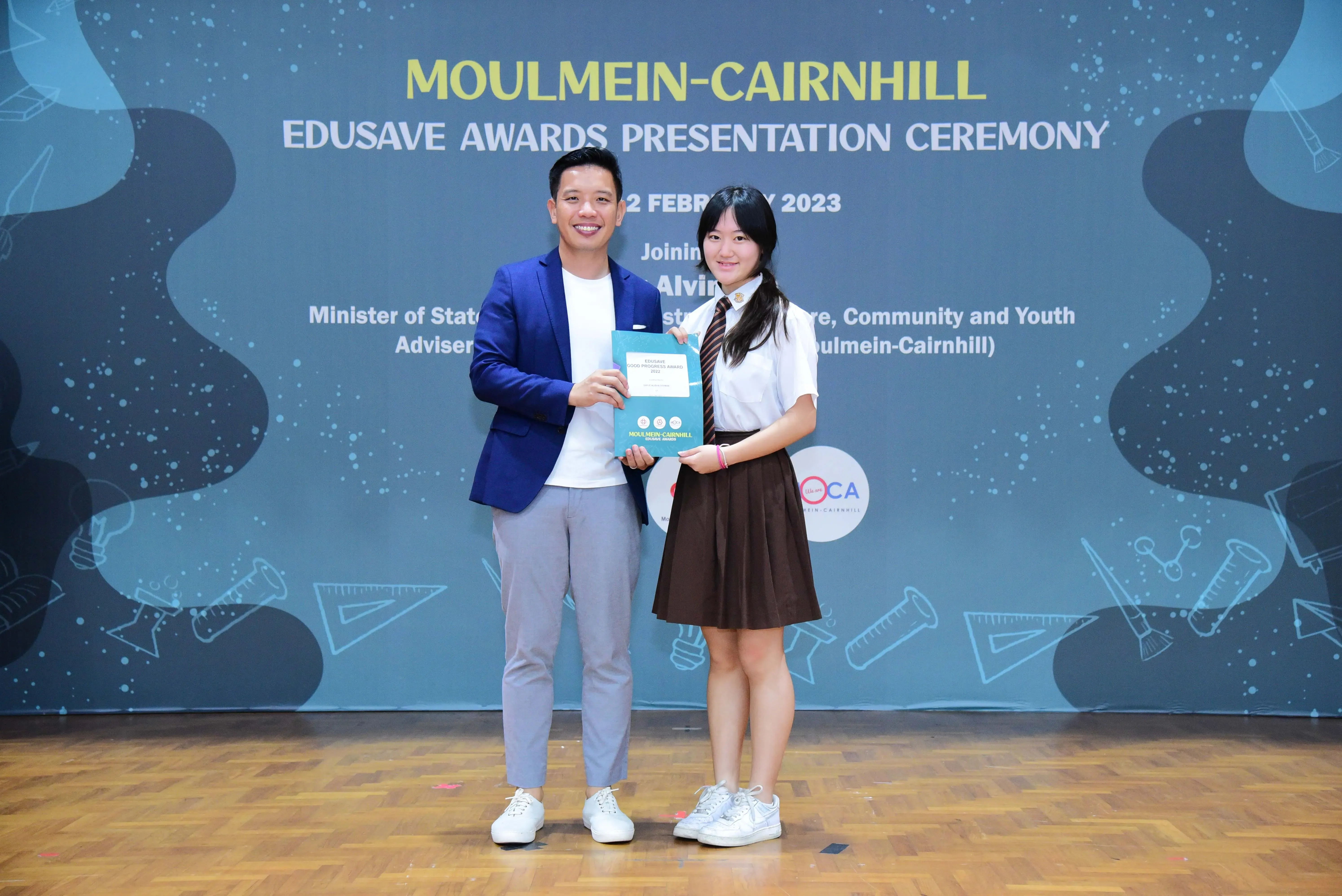 A man in a blue blazer and a female student in uniform hold a certificate at the Moulmein-Cairnhill Edusave Awards 2023.