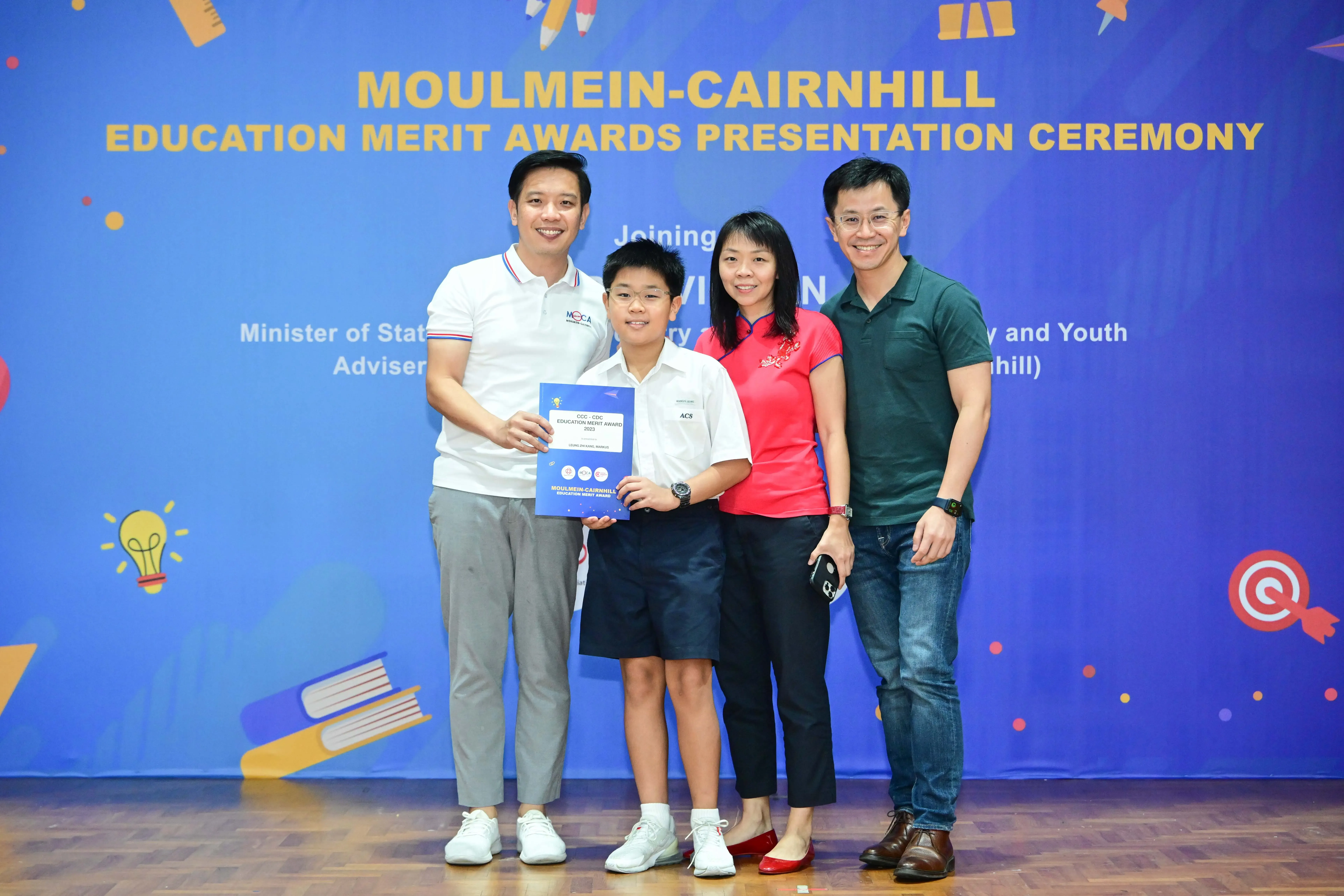 A boy holding a blue award certificate poses with three adults at the Moulmein-Cairnhill Education Merit Awards ceremony.