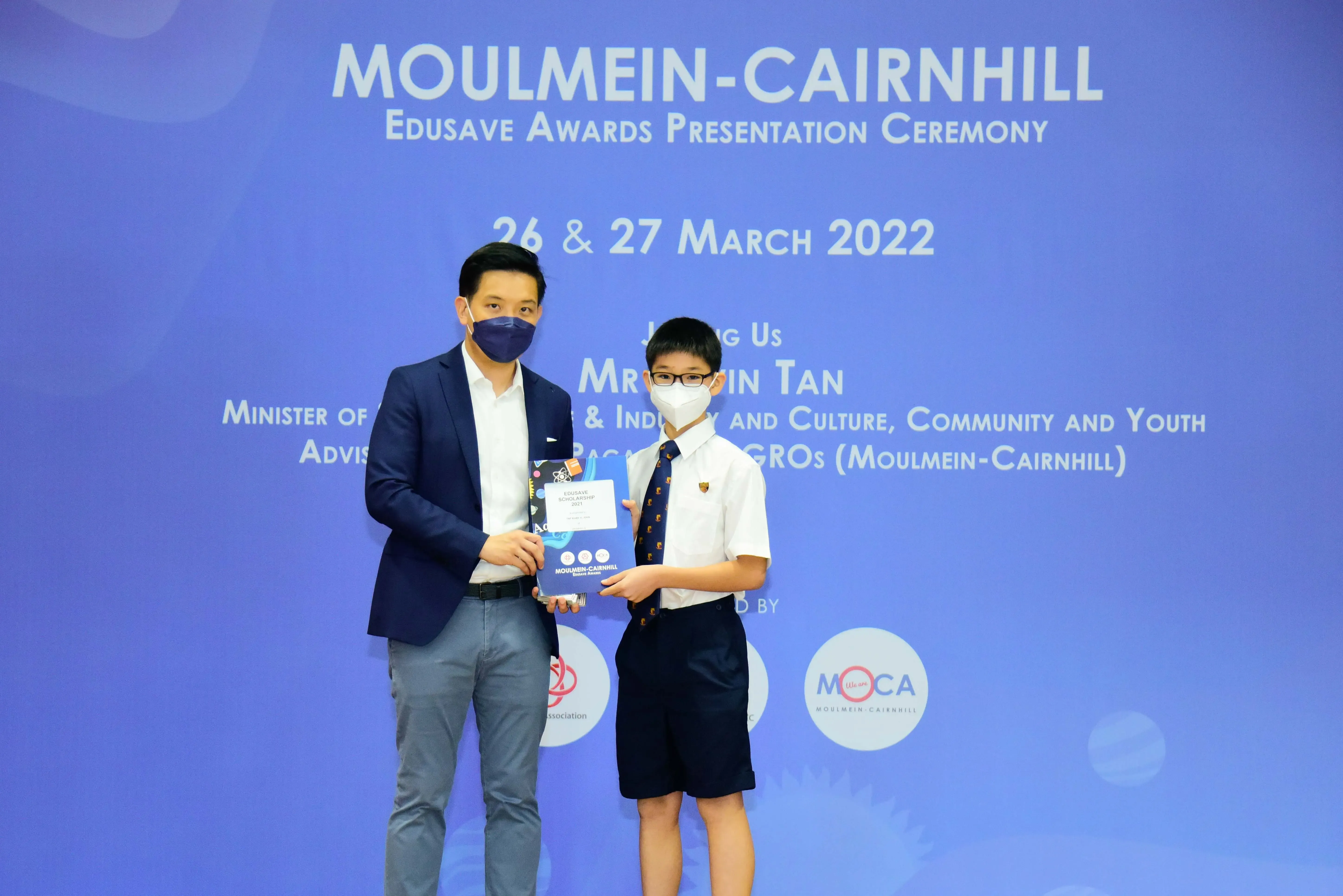 An adult and a young boy in school uniform hold an award at the Moulmein-Cairnhill Edusave Awards Ceremony 2022.