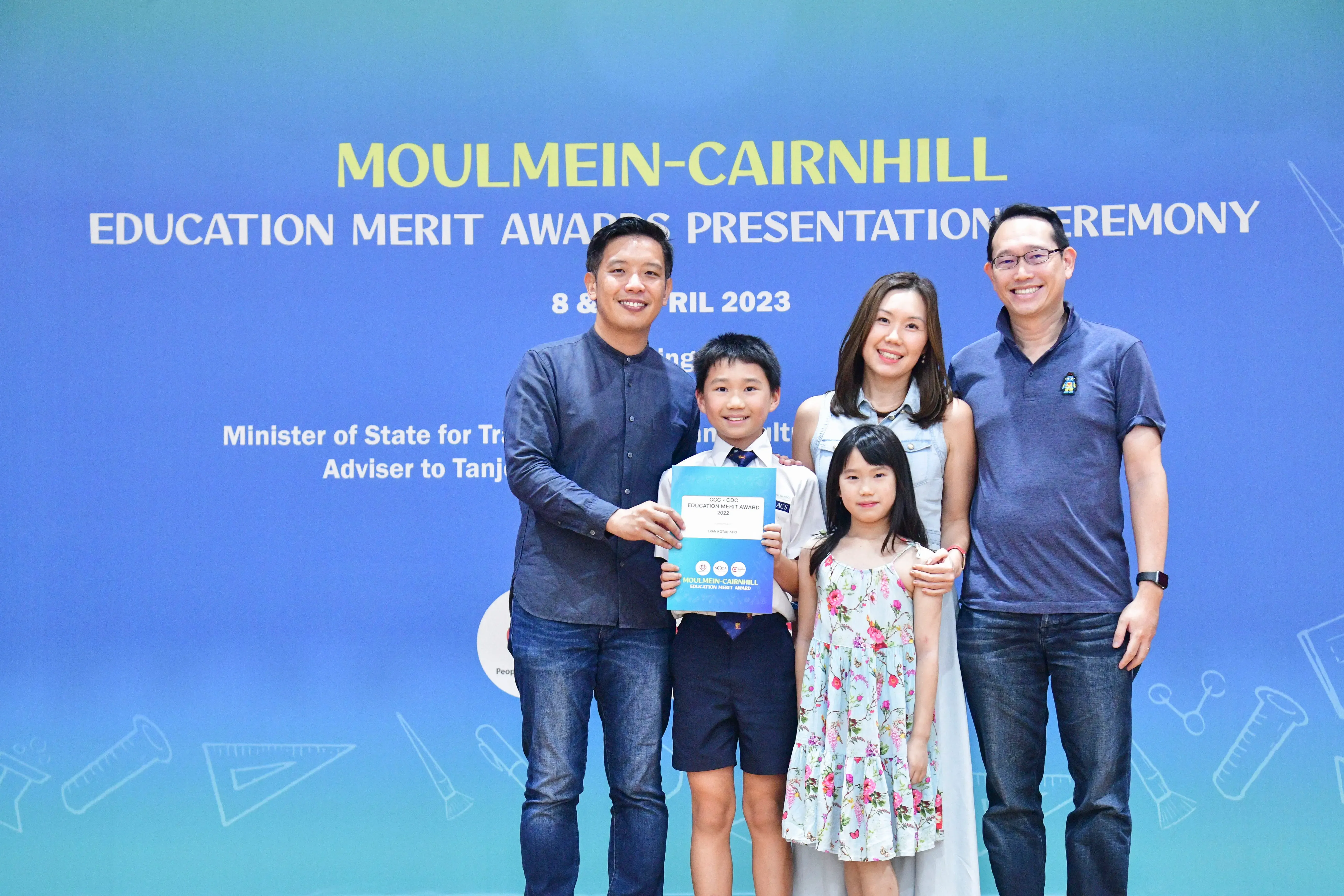 A boy holds an award certificate with four adults and a girl at the Moulmein-Cairnhill Education Merit Awards 2023.