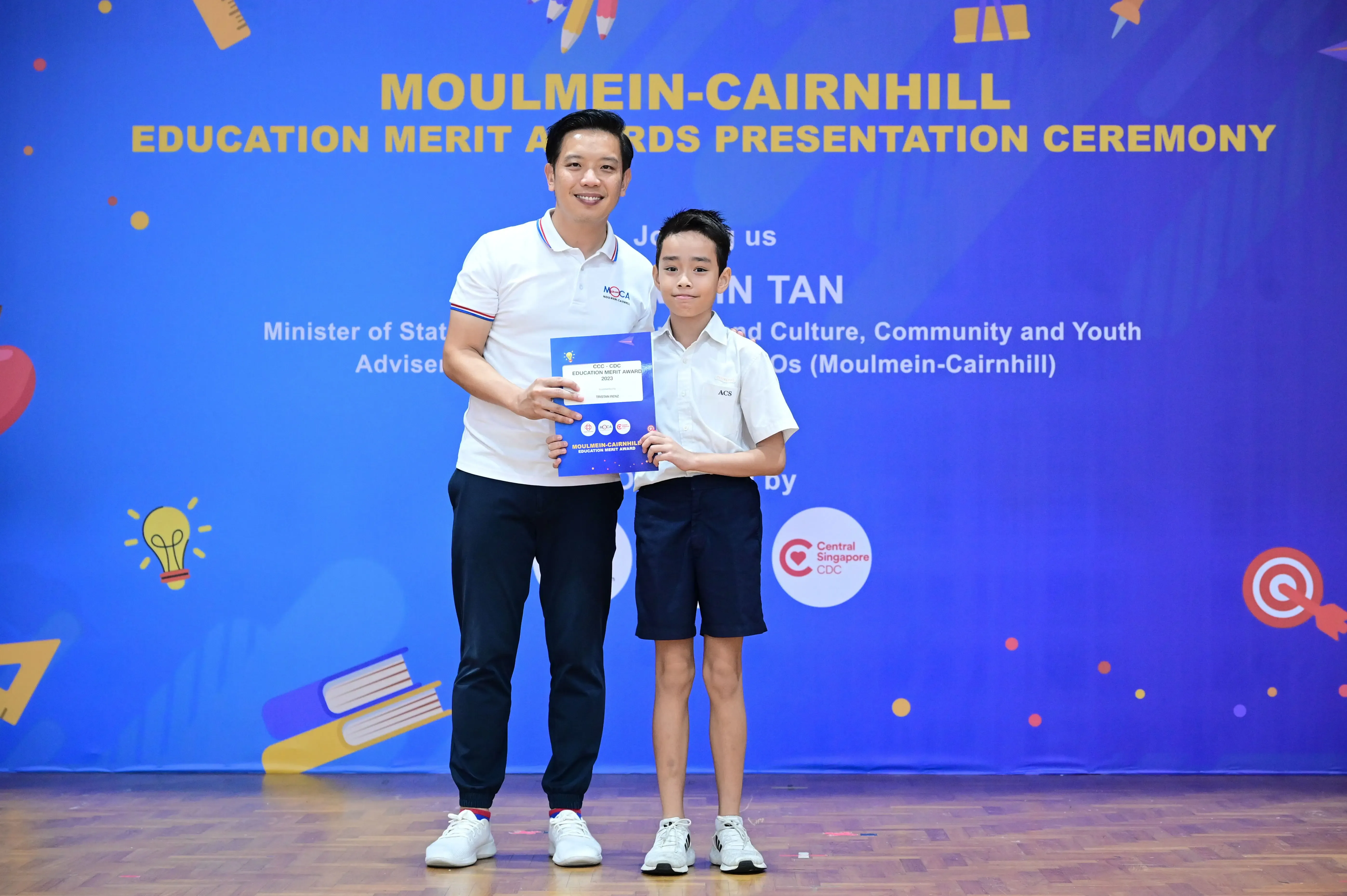 An adult and a young boy hold a certificate together at the Moulmein-Cairnhill Education Merit Awards Presentation Ceremony.