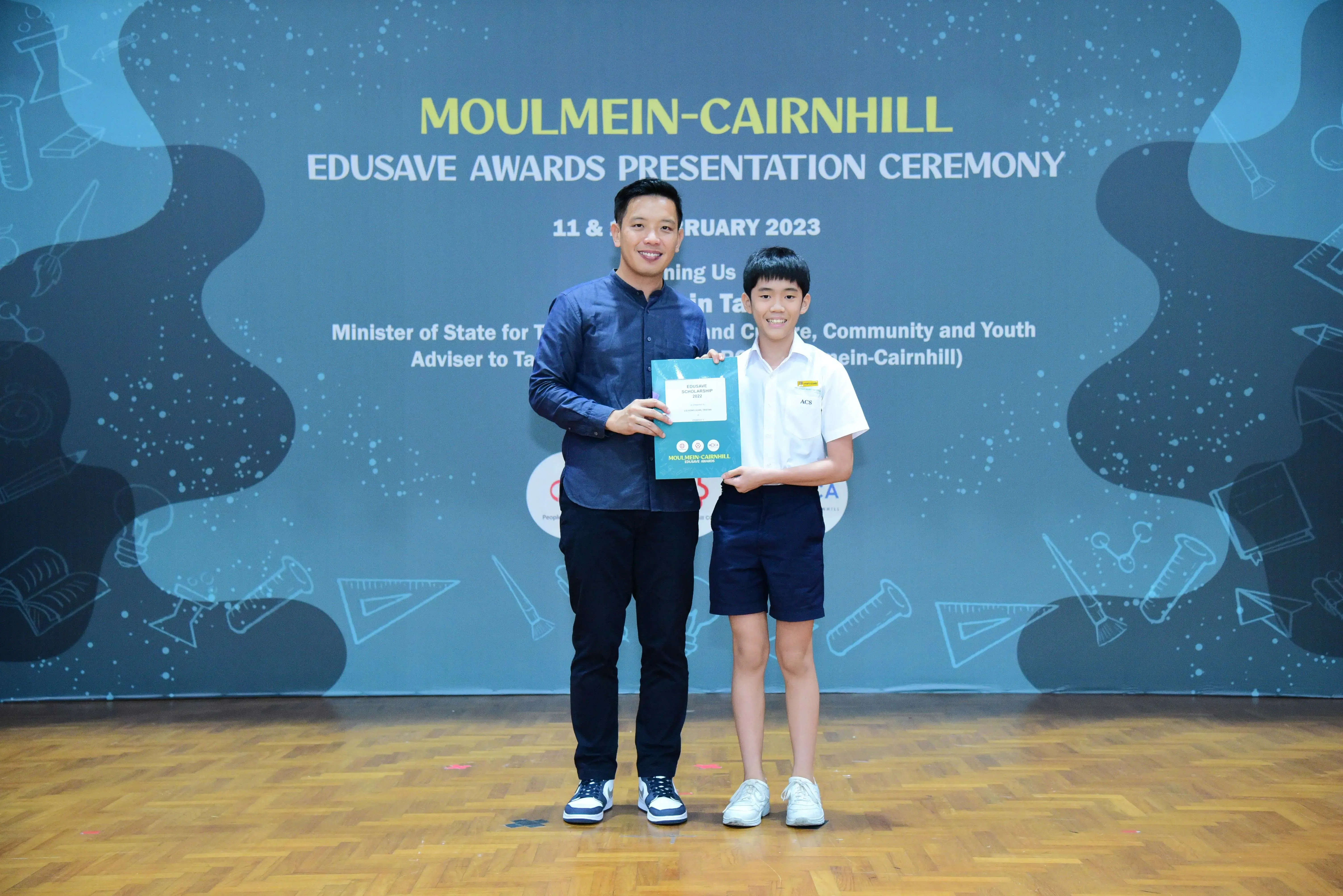 An adult and a young boy hold a teal certificate together at the Moulmein-Cairnhill Edusave Awards Ceremony 2023.