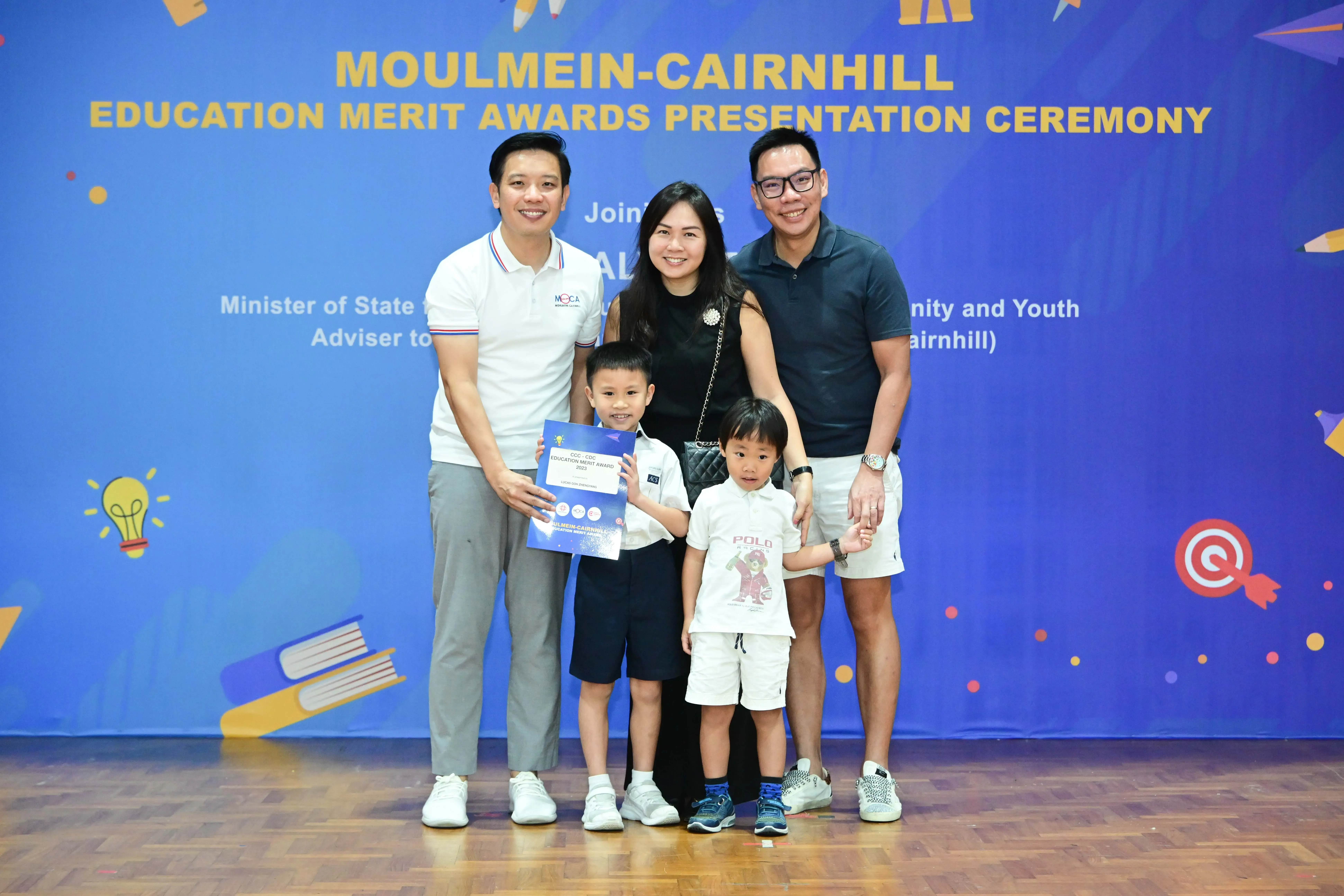 Three adults and two young boys pose smiling before a Moulmein-Cairnhill Education Merit Awards banner, one boy holding a certificate.