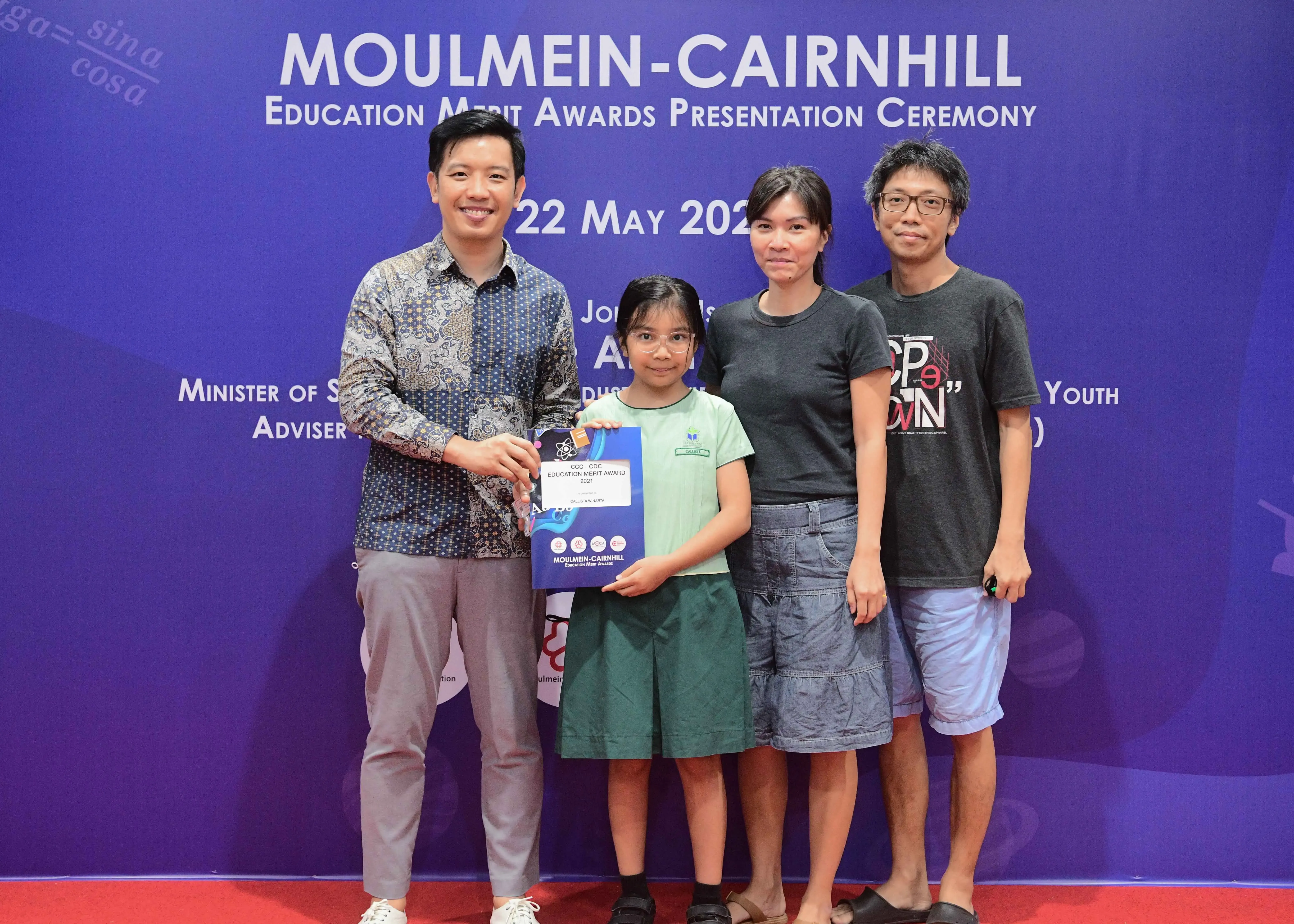 Four people pose at the Moulmein-Cairnhill Education Merit Awards ceremony, with a girl holding an award certificate.
