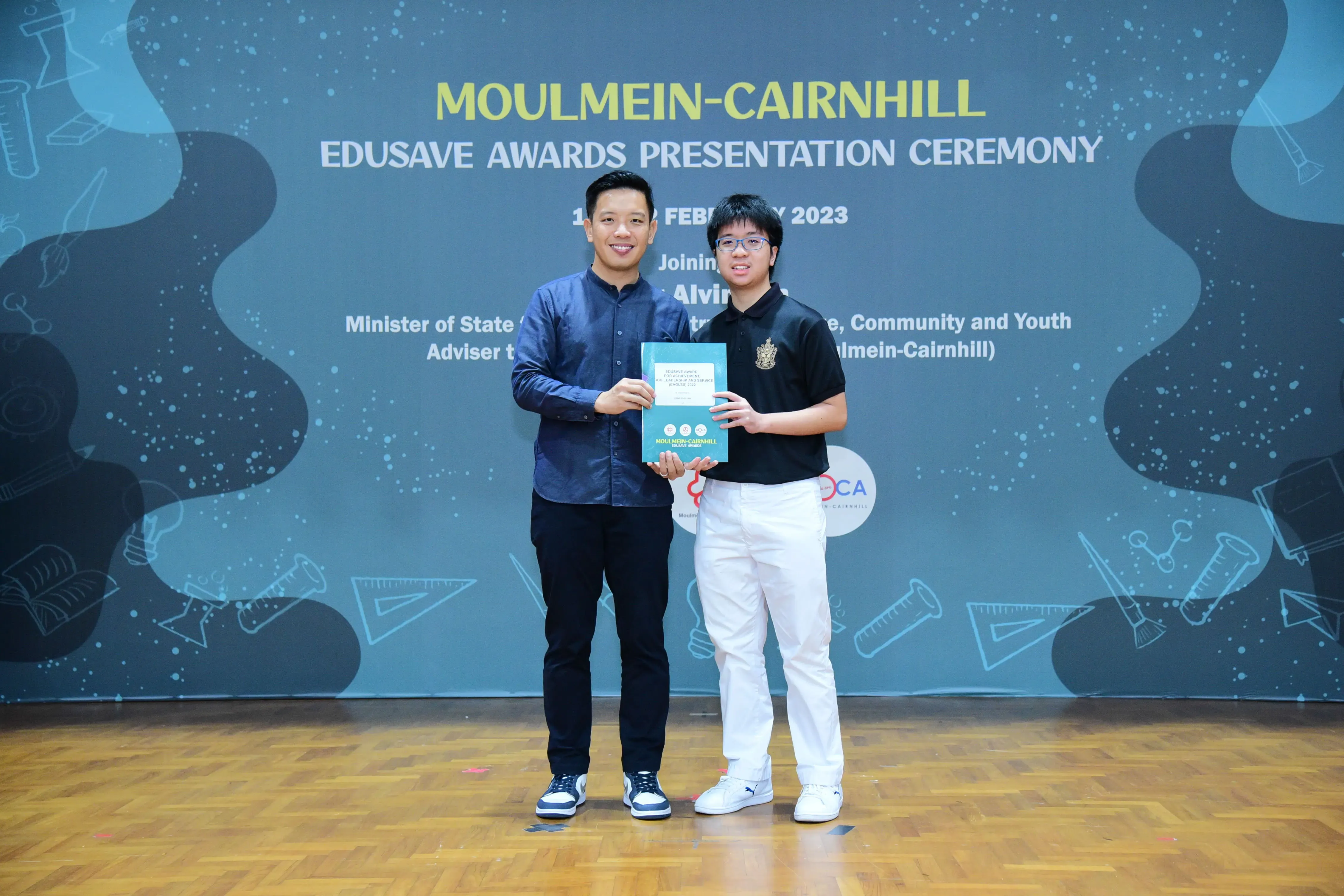 Two people smile while holding a certificate at the Moulmein-Cairnhill Edusave Awards Presentation Ceremony 2023.