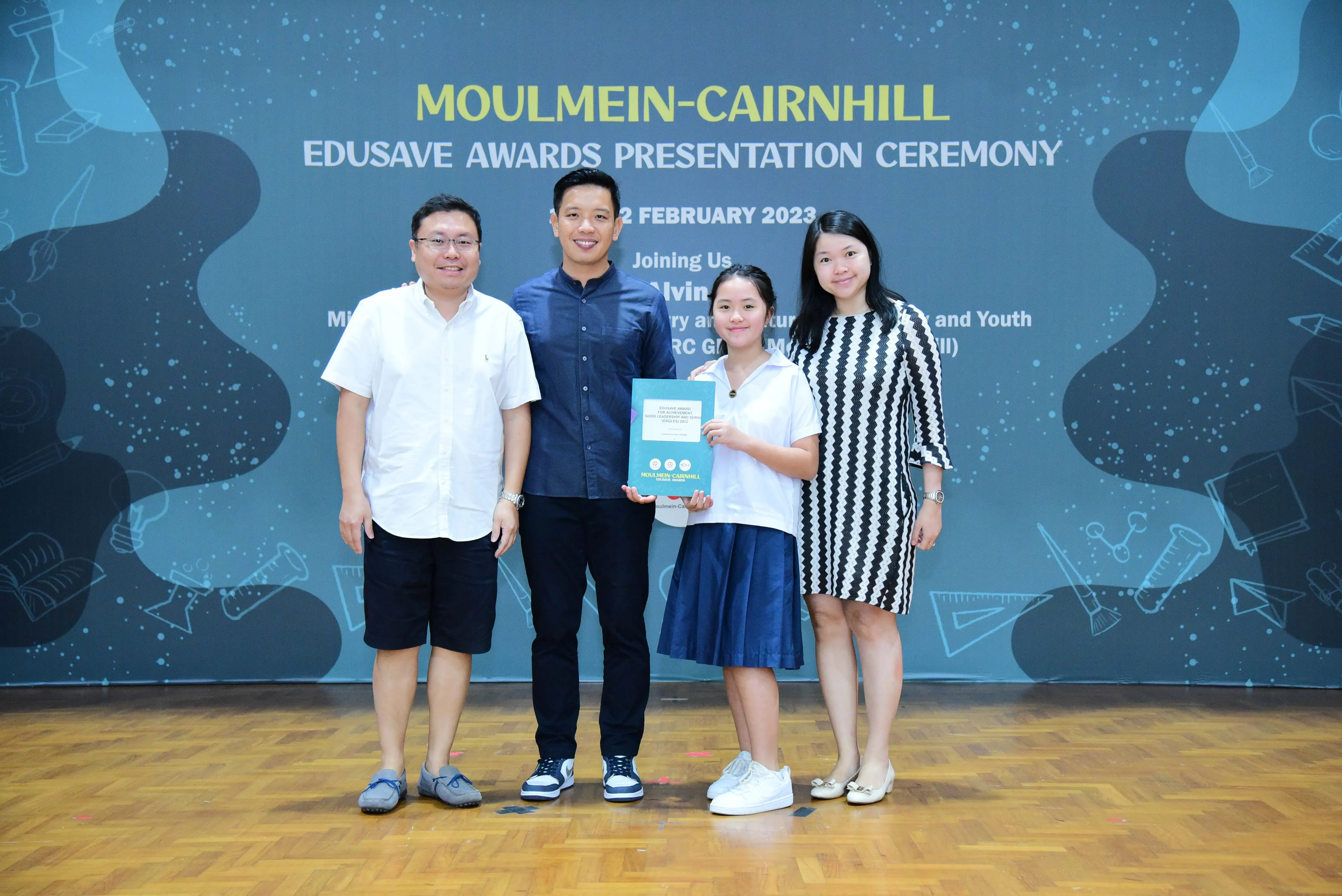 Four people pose at the Moulmein-Cairnhill Edusave Awards Presentation Ceremony, with a student holding an award booklet.