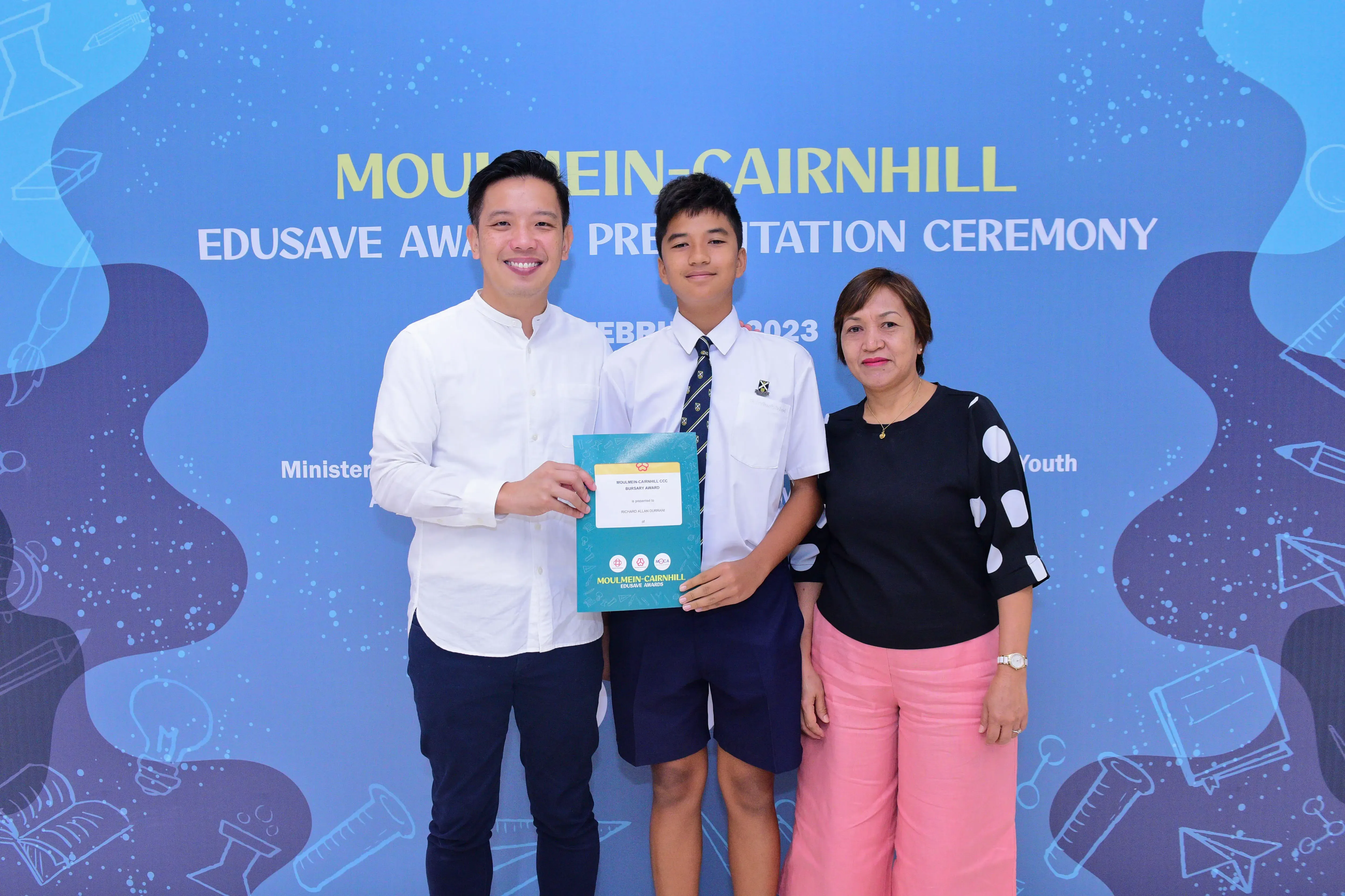 A student in school uniform holds a certificate flanked by two adults at the Moulmein-Cairnhill Edusave Award Presentation Ceremony 2023.